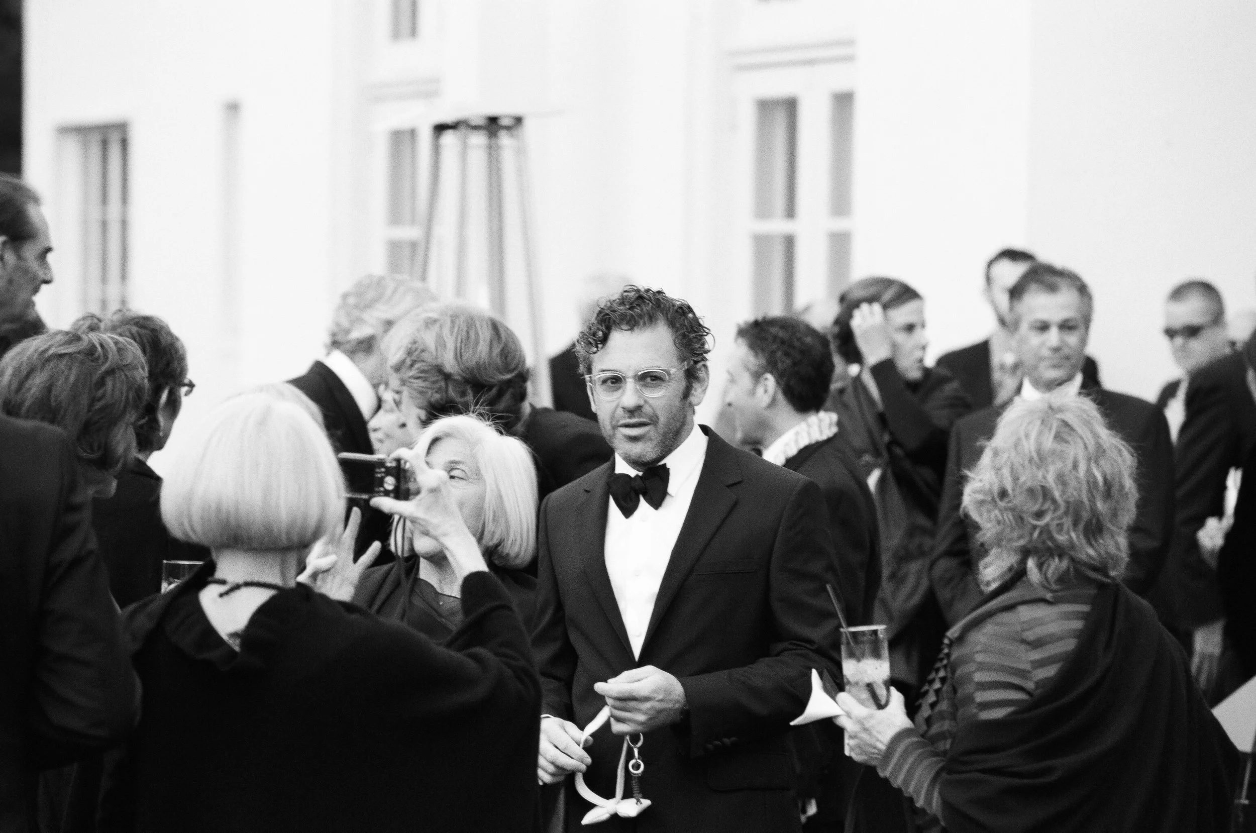 A man in a black tuxedo with a bowtie, wearing glasses, is standing amidst a group of people, many of whom are taking photos or videos. The scene appears to be at a formal event or gathering in a spacious, well-lit room with high ceilings and large windows.