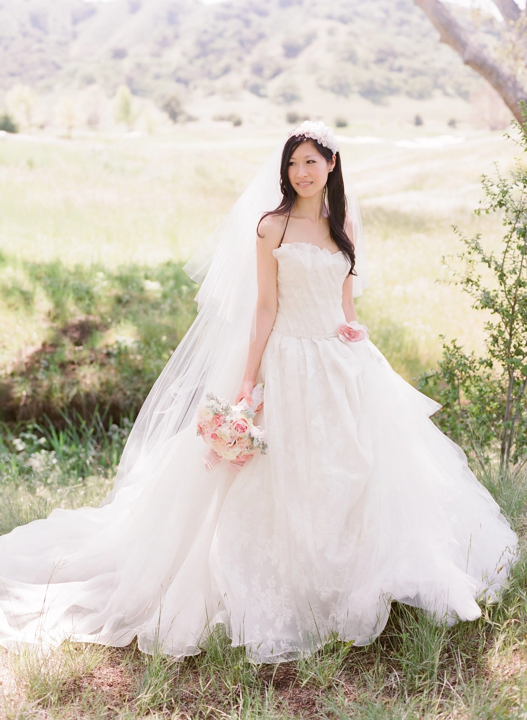 A bride in a white wedding gown standing outdoors in a grassy area with trees and hills in the background. She holds a bouquet of pink and white flowers and wears a floral headpiece.