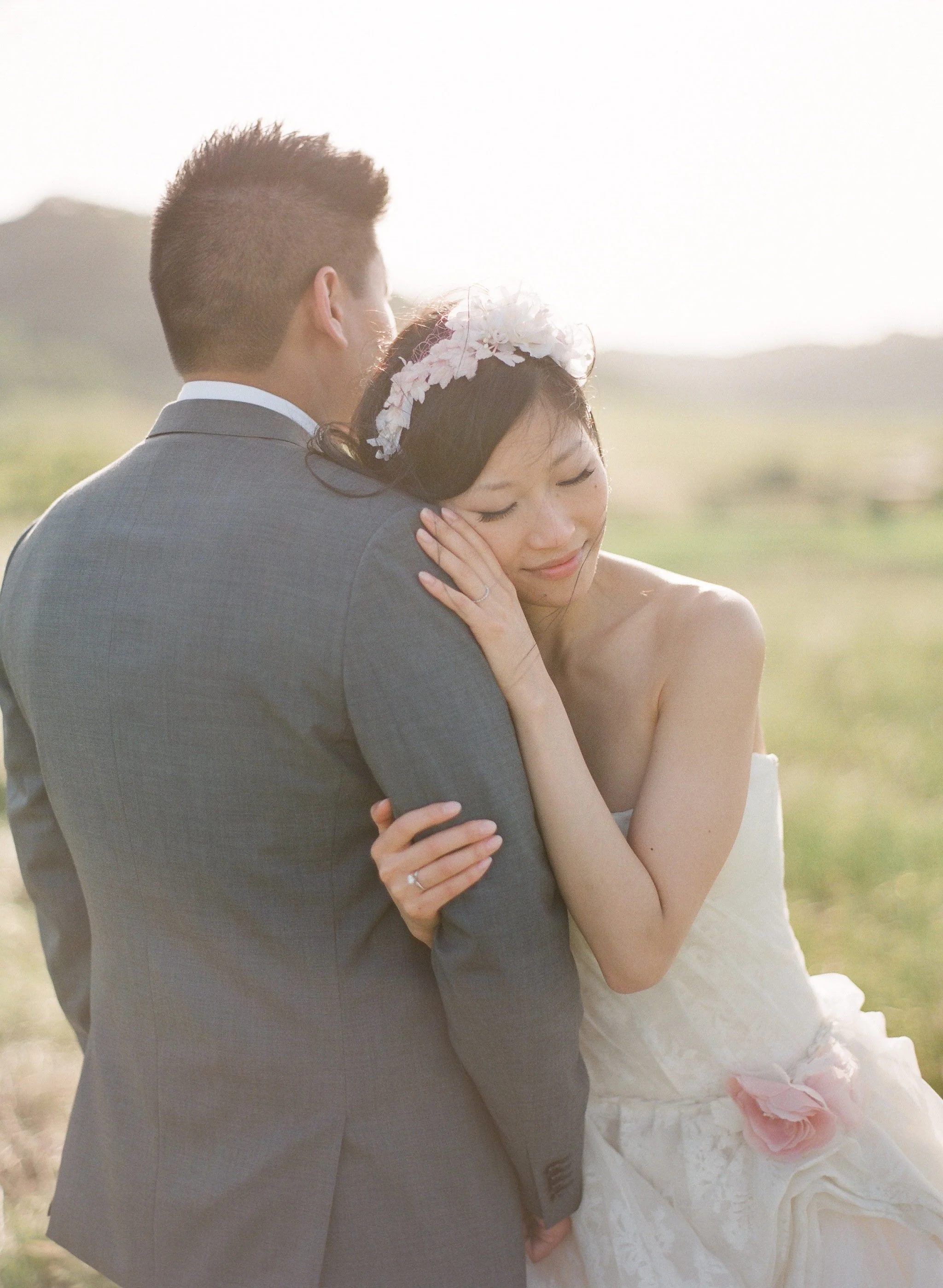 A bride and groom embrace outdoors in a picturesque landscape, showcasing wedding attire and a tender moment.