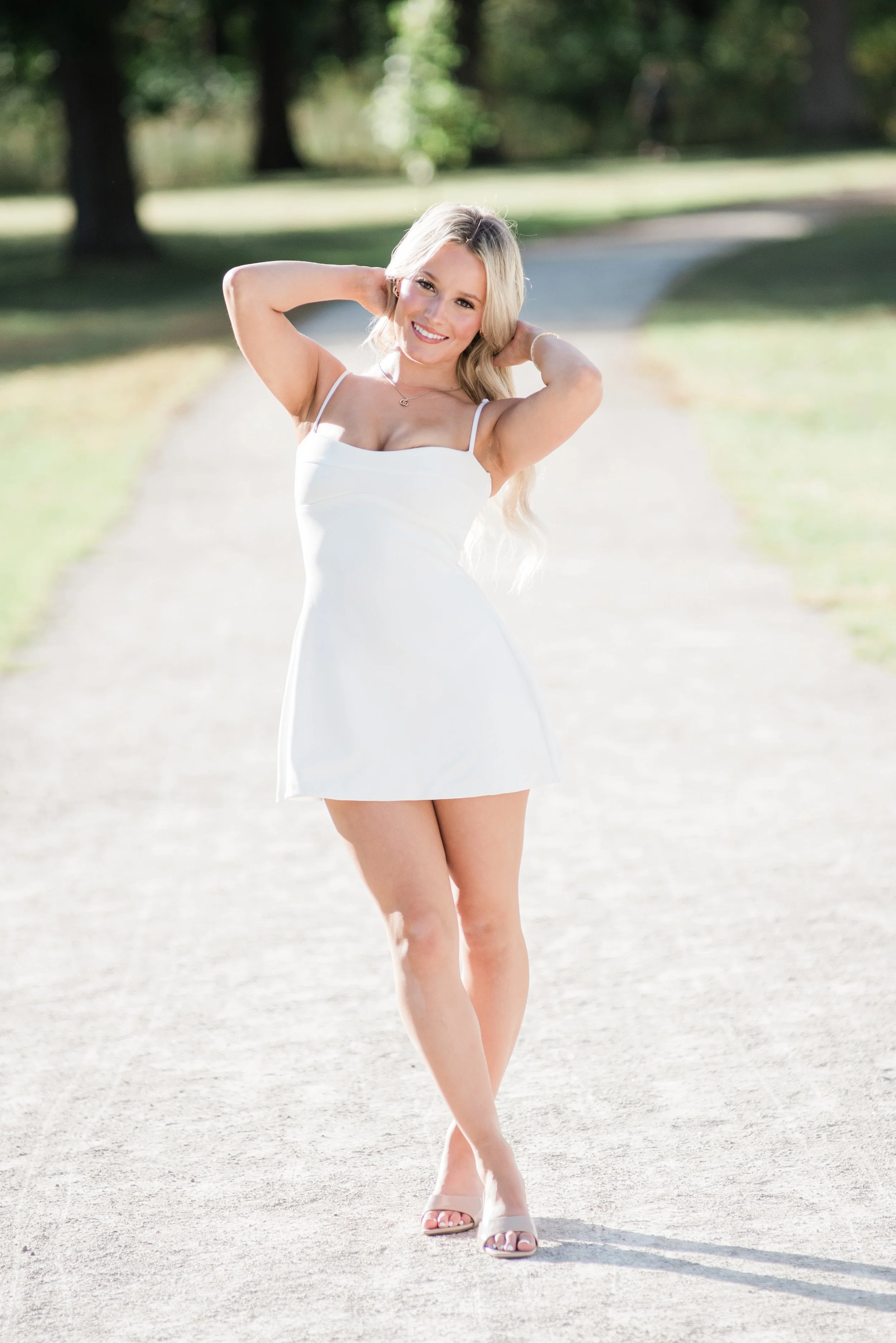 A young woman with blonde hair in a white dress standing outdoors on a gravel path, smiling with her hands behind her head, in a park-like setting with trees in the background.
