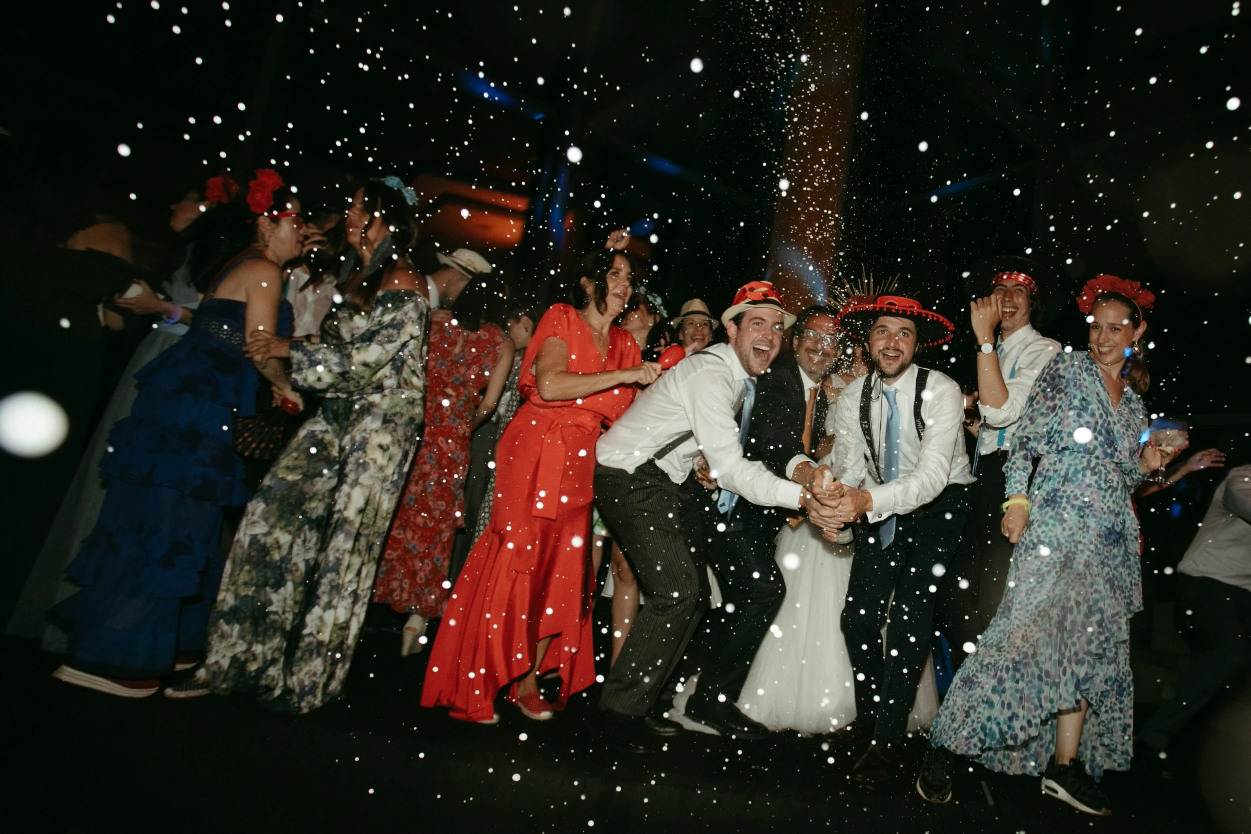 Grupo de personas en la fiesta, algunas con vestimentas tradicionales y accesorios como sombreros y coronas de flores, celebrando y lanzando confeti en un ambiente festivo.
