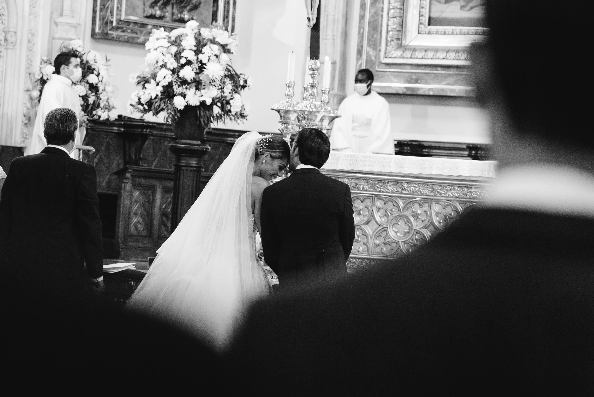 Una pareja en su boda, con la novia usando un vestido de novia y un velo, y el novio con traje oscuro, en una ceremonia en una iglesia antigua decorada con flores y candelabros.