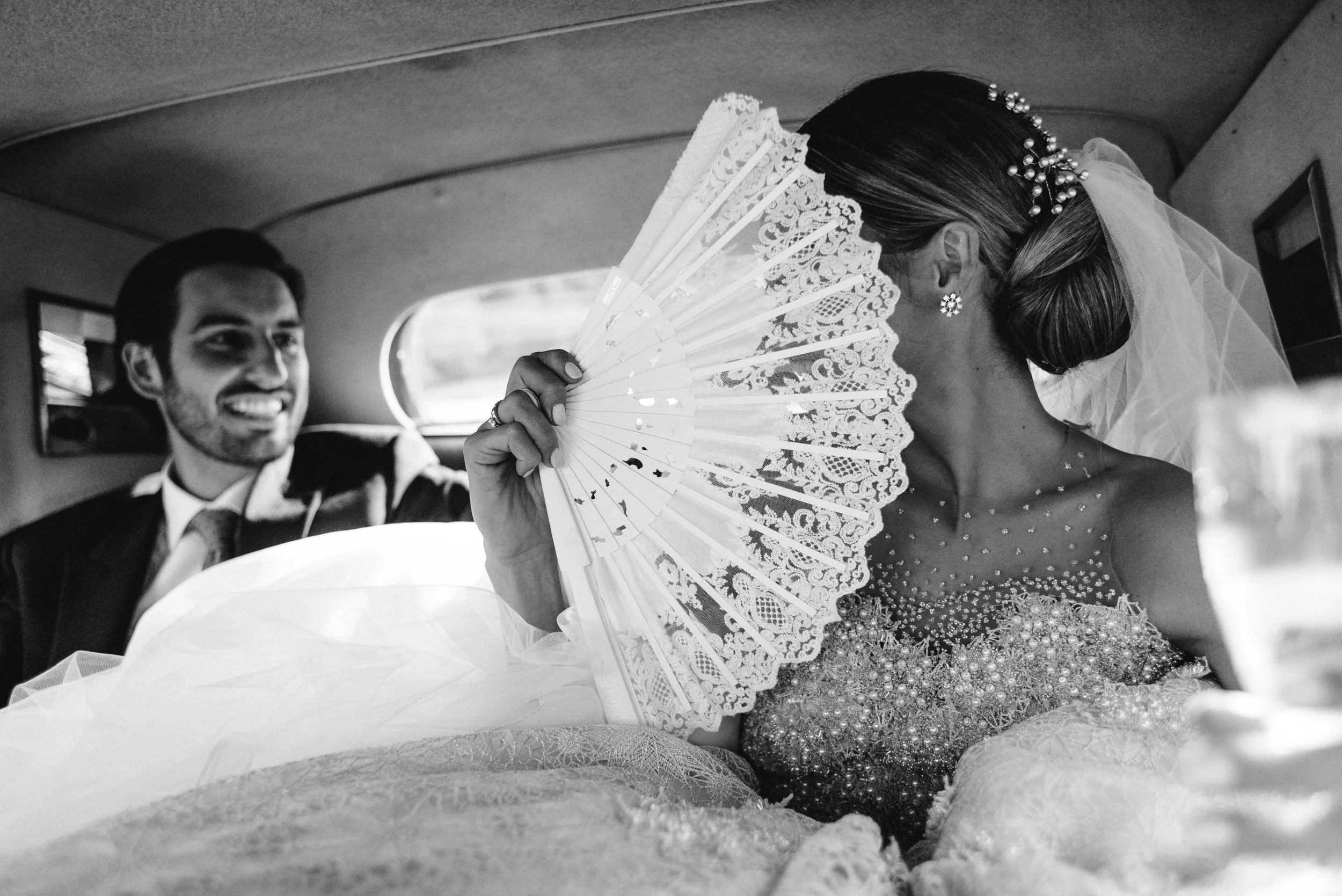 Una pareja en un coche de boda, la mujer viste un vestido con detalles brillantes y lleva un velo, ella sostiene un abanico de encaje frente a su rostro, y el hombre sonríe mientras la mira.
