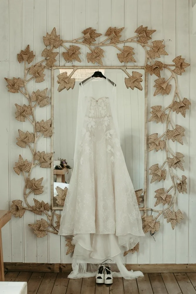 Vestido de novia colgado en un marco decorado con hojas de yuca. Debajo del vestido hay unos zapatos blancos en el suelo.