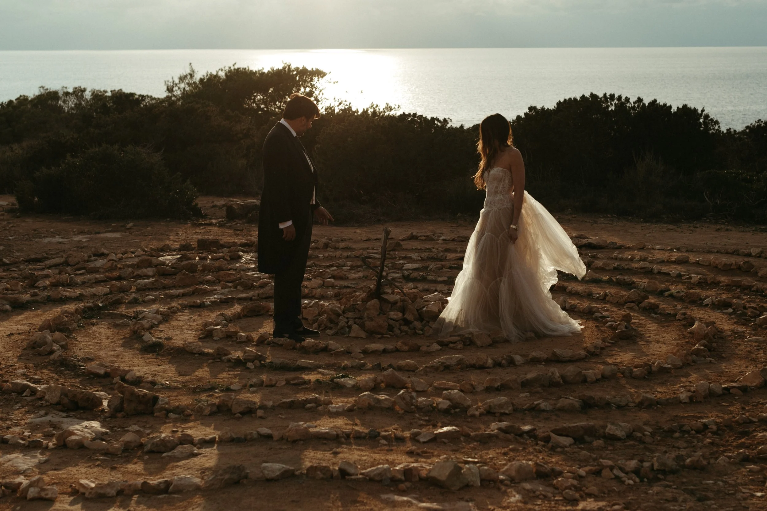 Pareja de novios en ceremonia en un entorno natural al atardecer, con escenario de rocas y vegetación alrededor del mar.