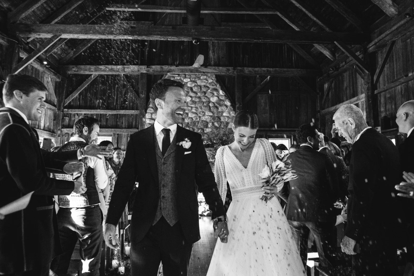 Una pareja de recién casados sonrientes caminando entre invitados en una ceremonia de boda en un espacio rústico con estructura de madera y chimenea de piedra.