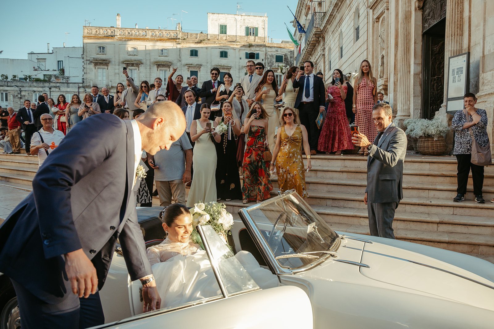 The couple leaving in a classic car in front of steps full of guests