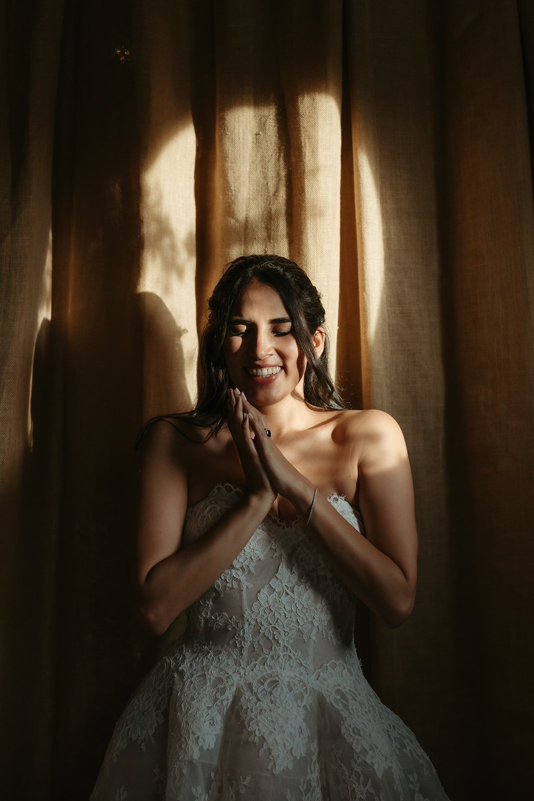 Camila sonríe con las manos juntas a la luz de una cortina