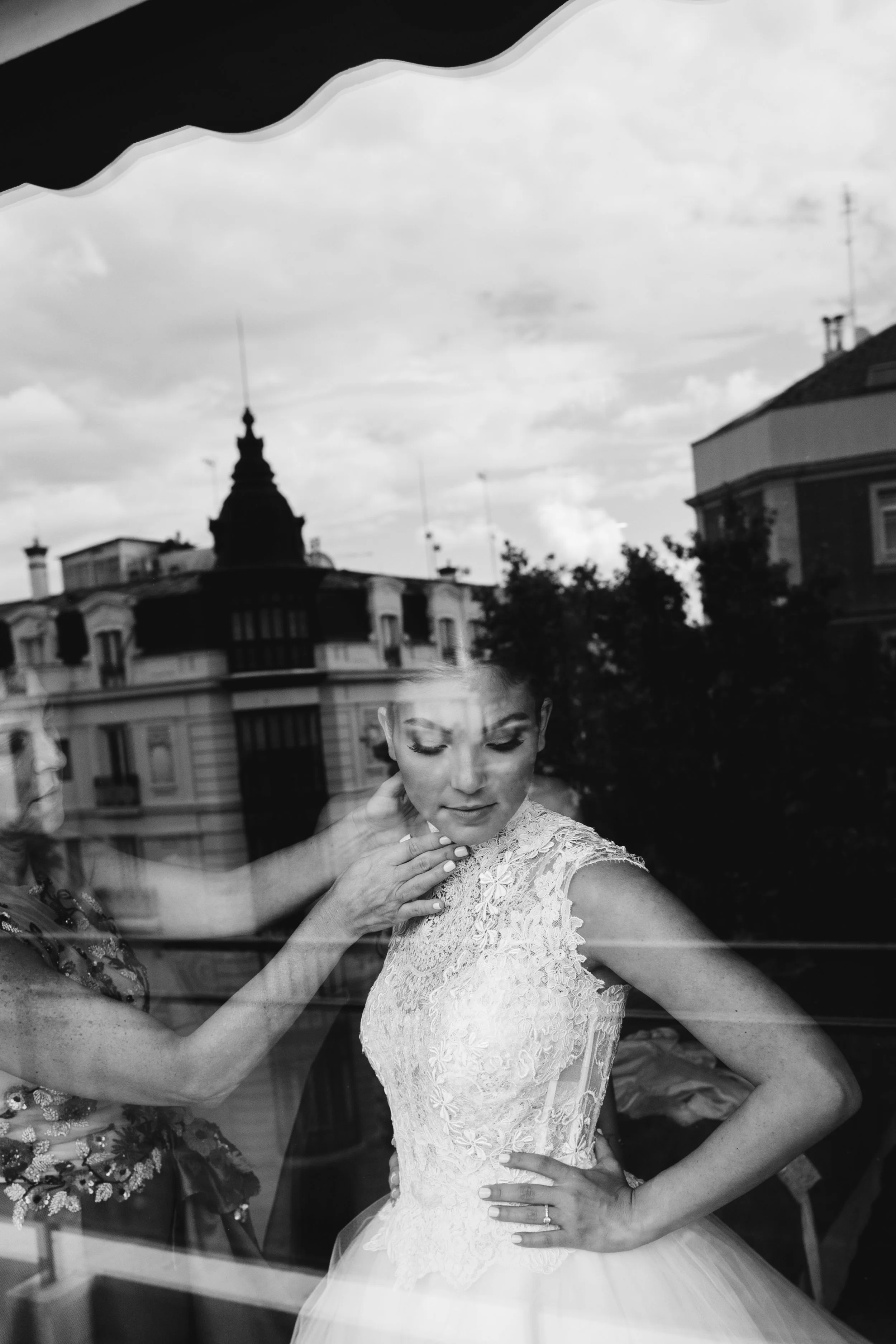 Mujer con vestido de novia siendo arreglada, vista a través de una ventana, con reflejo de edificios en el cristal, en blanco y negro.