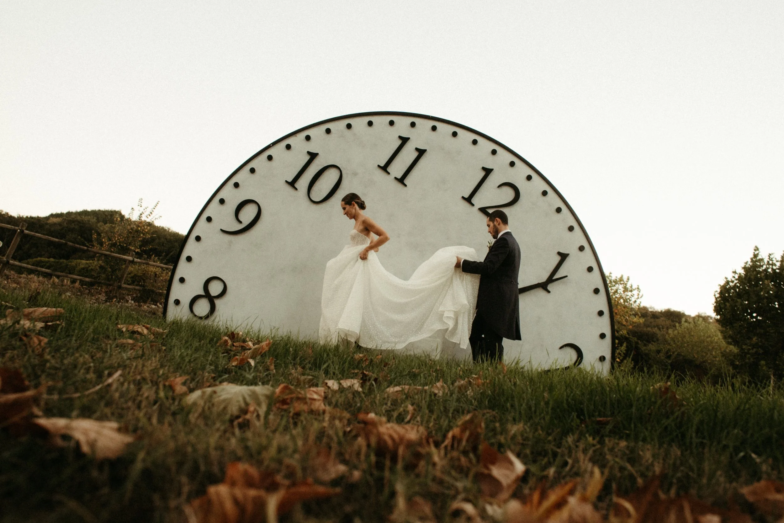 Pareja de novios en una escena de boda romántica frente a un gran reloj al aire libre, con árboles en el fondo y hojas en el suelo.