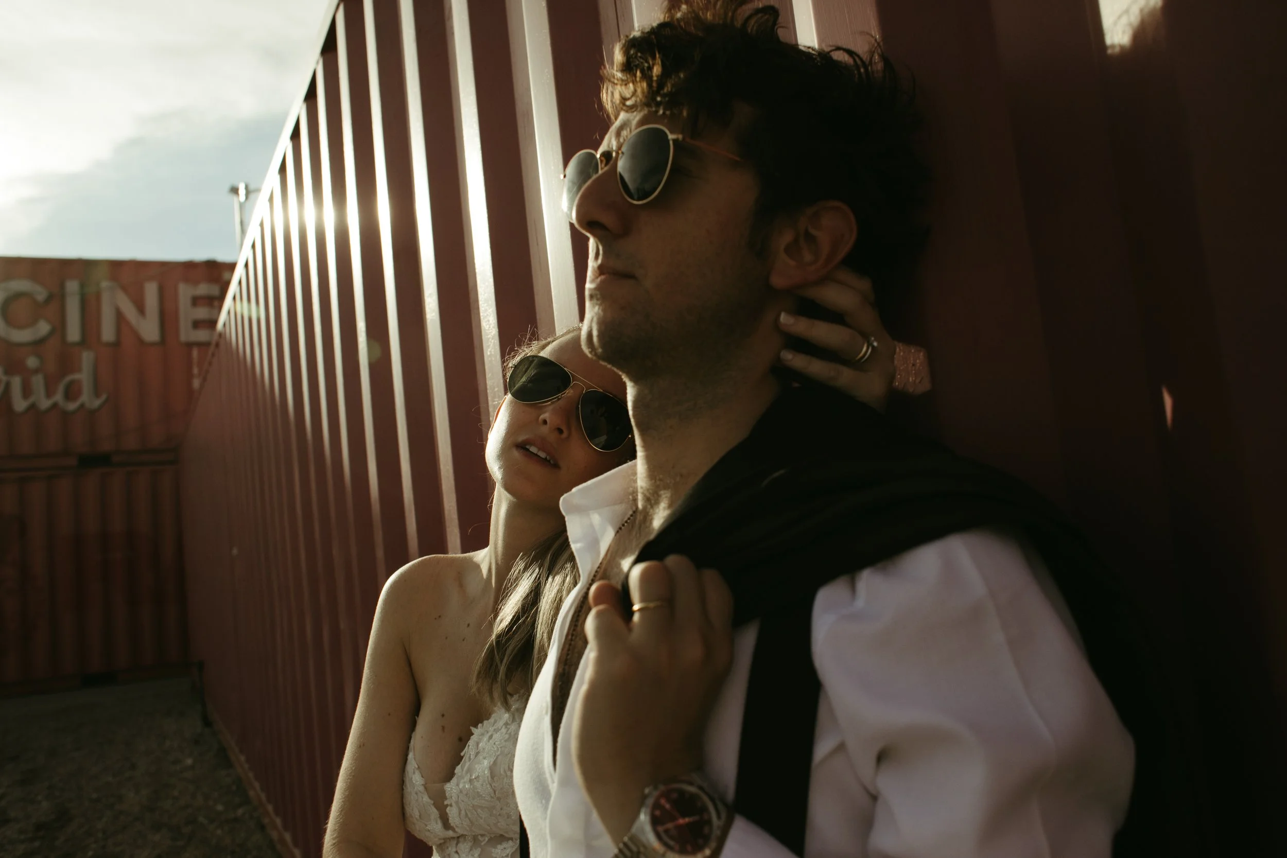 Una pareja usando gafas de sol, apoyada contra una cerca roja en un entorno al aire libre con nubes y un cartel que dice 'CINE' en el fondo.