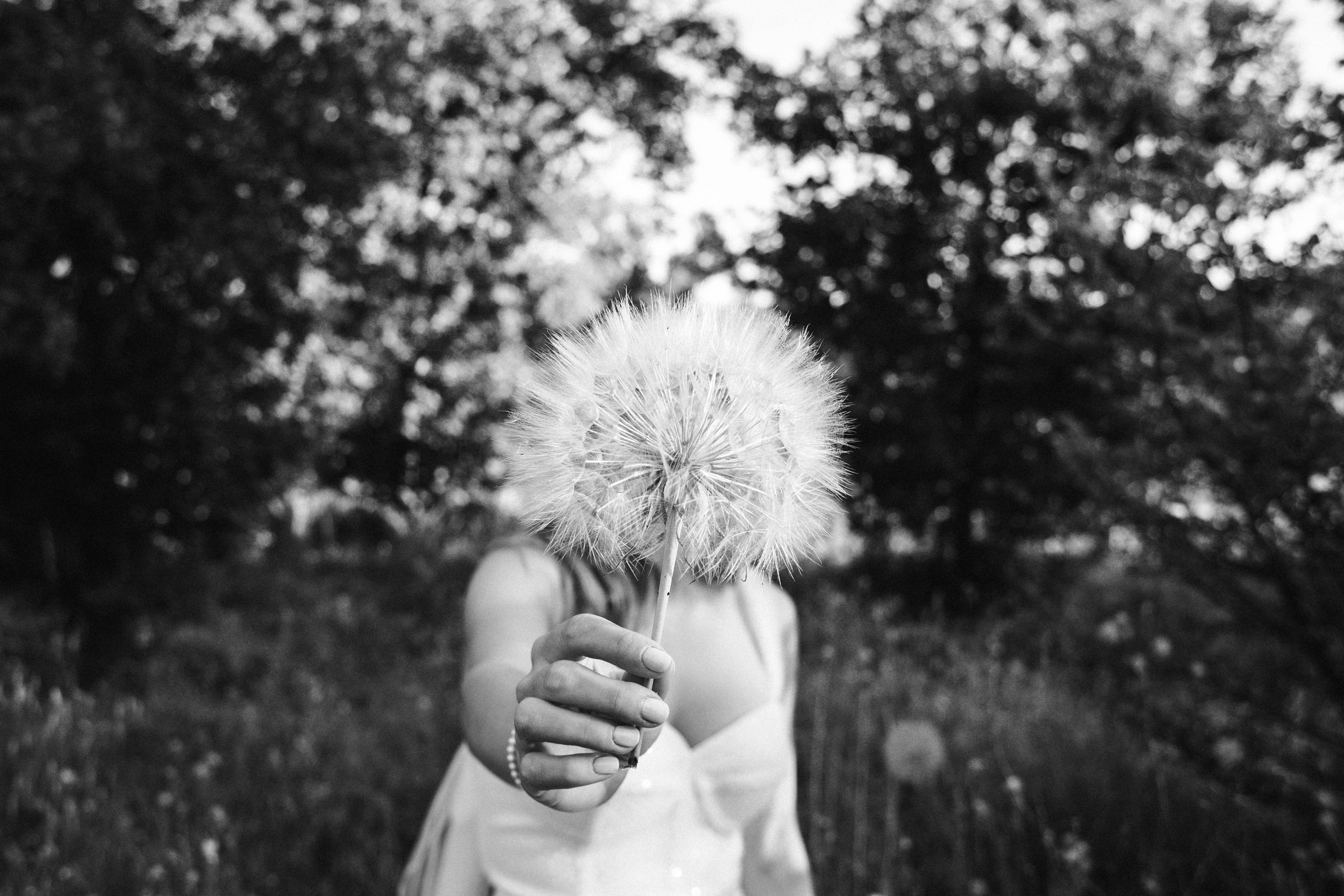 Persona sosteniendo un diente de león en un entorno natural con árboles en el fondo, en una imagen en blanco y negro.