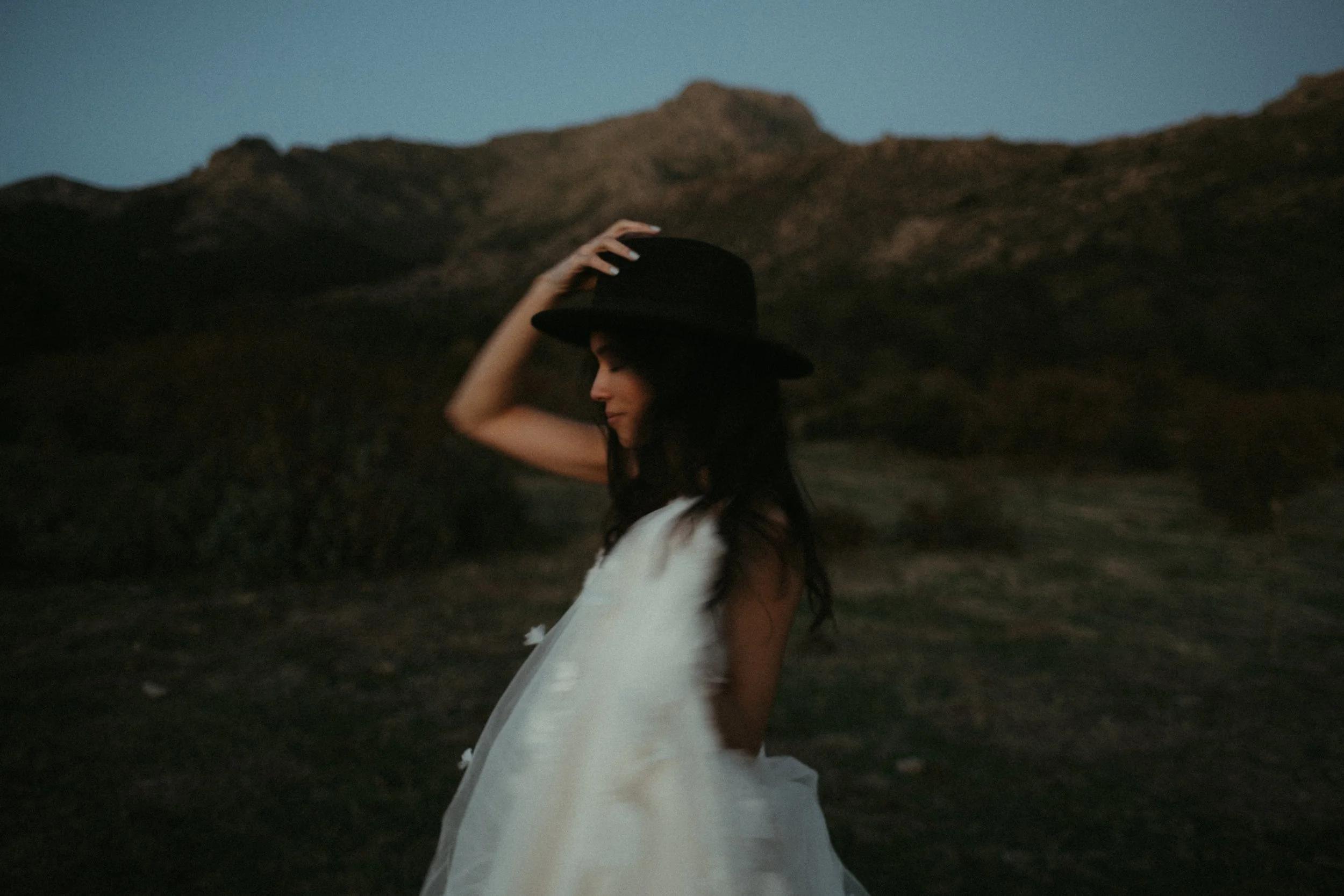 Mujer con vestido blanco y sombrero negro en tierra árida con montañas al atardecer