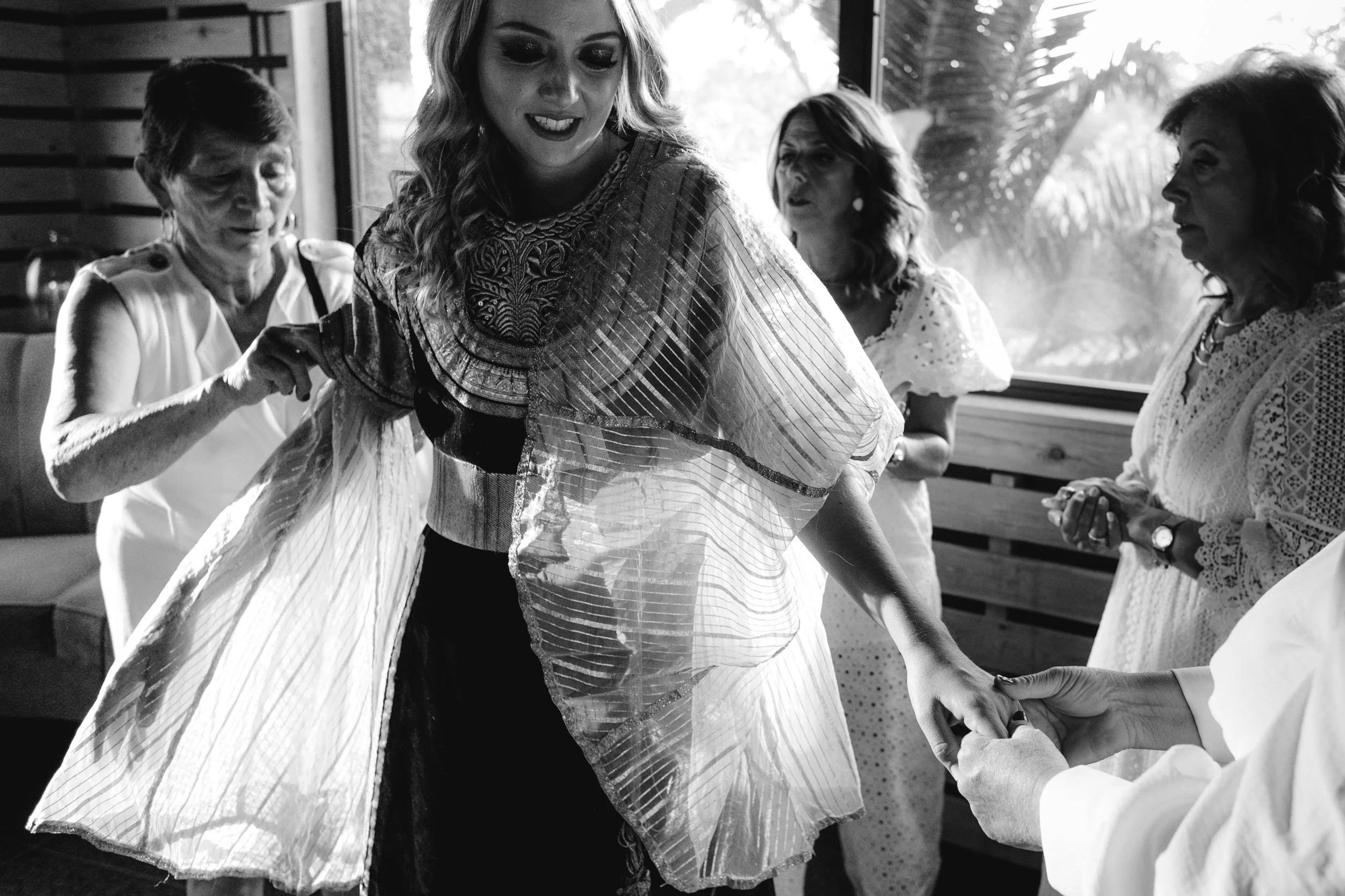 Mujer sonriendo mientras toma la mano de otra persona en un ambiente interior, acompañada por varias mujeres observando y participando en la escena.