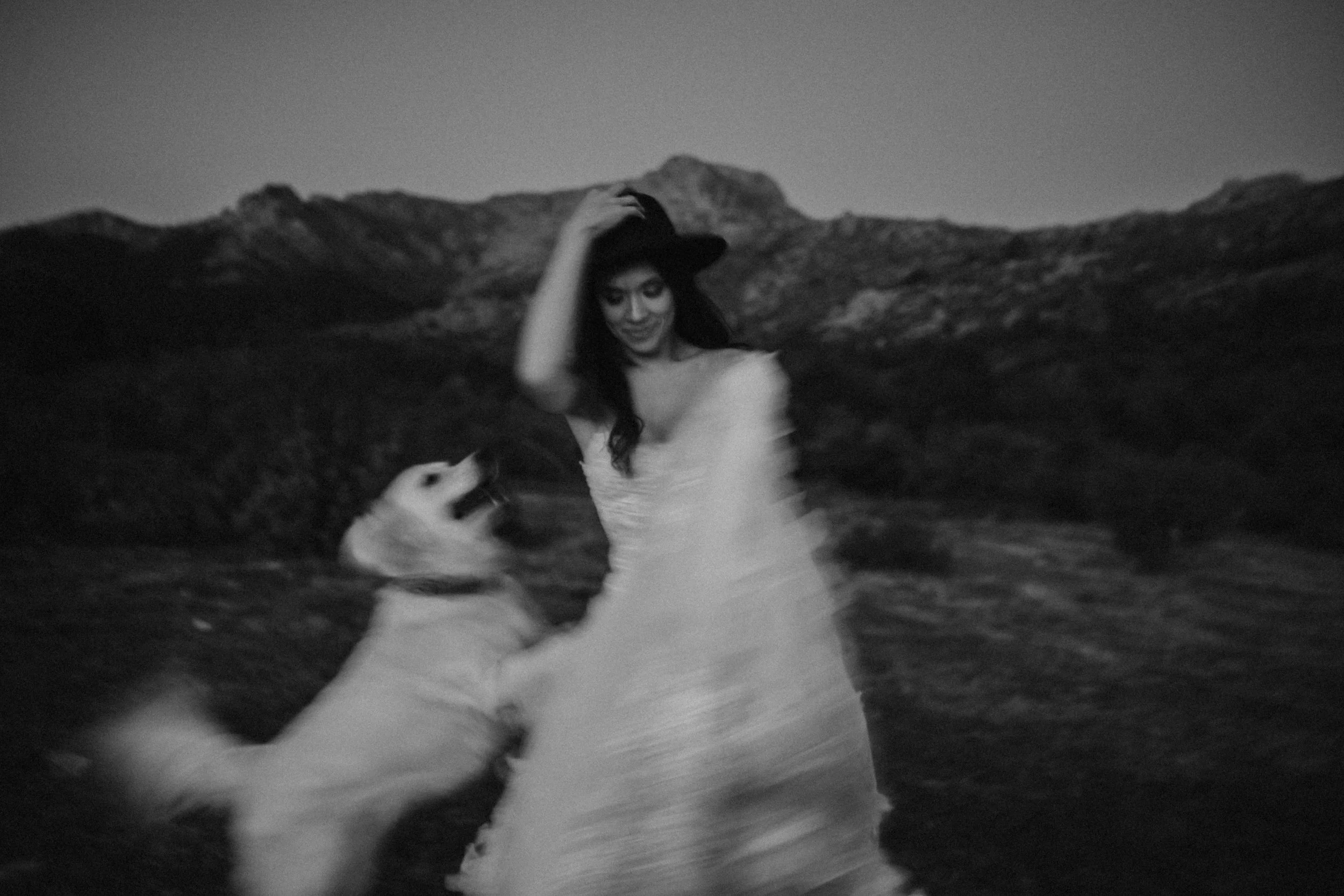 Una mujer vestida con un vestido largo y un sombrero, sonriendo, en un paisaje montañoso al atardecer, con una persona al lado que la sostiene por la espalda.