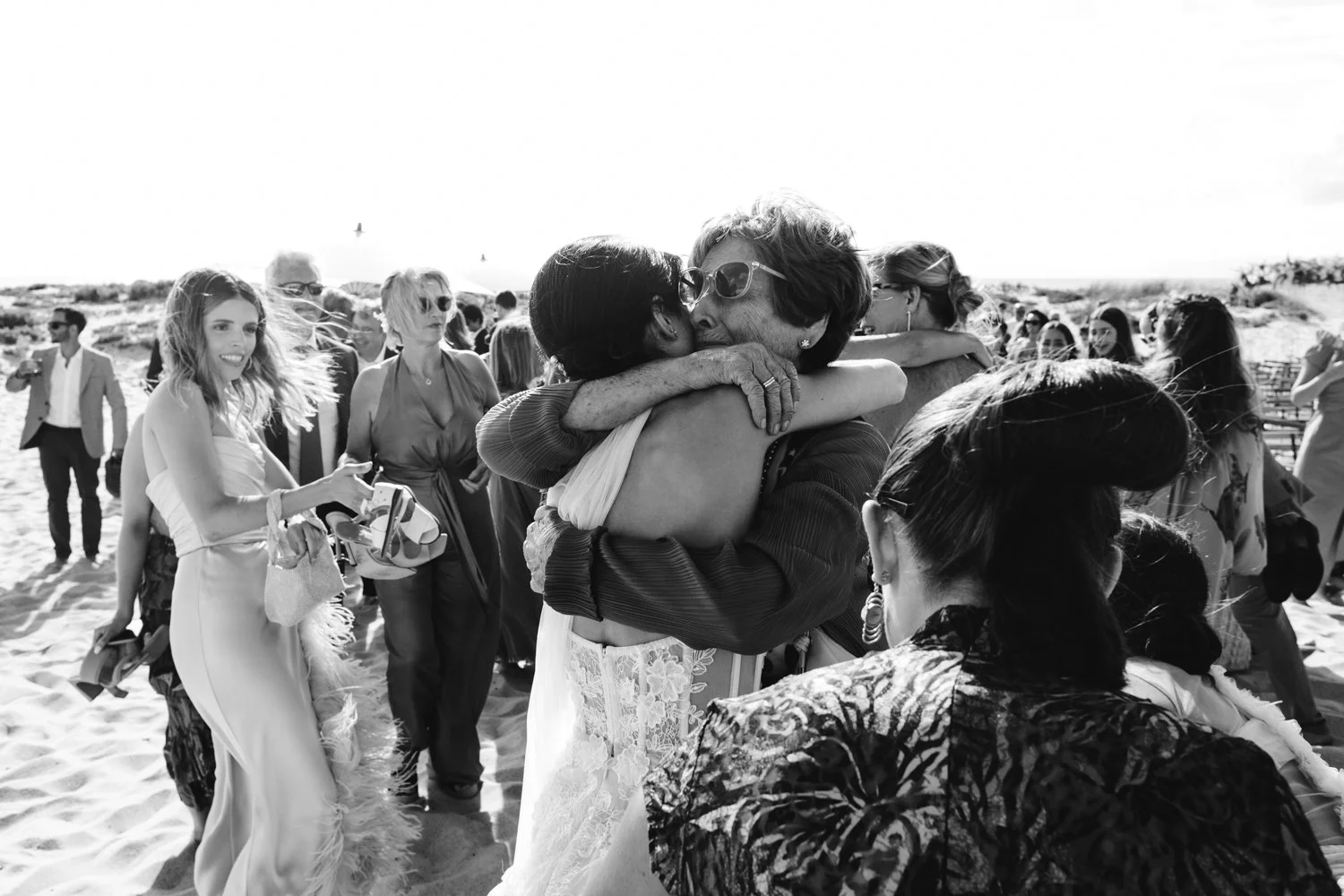 Dos mujeres se abrazan y se besan en la playa durante una reunión social, rodeadas de varias personas que observan y participan en la celebración, en un día soleado.