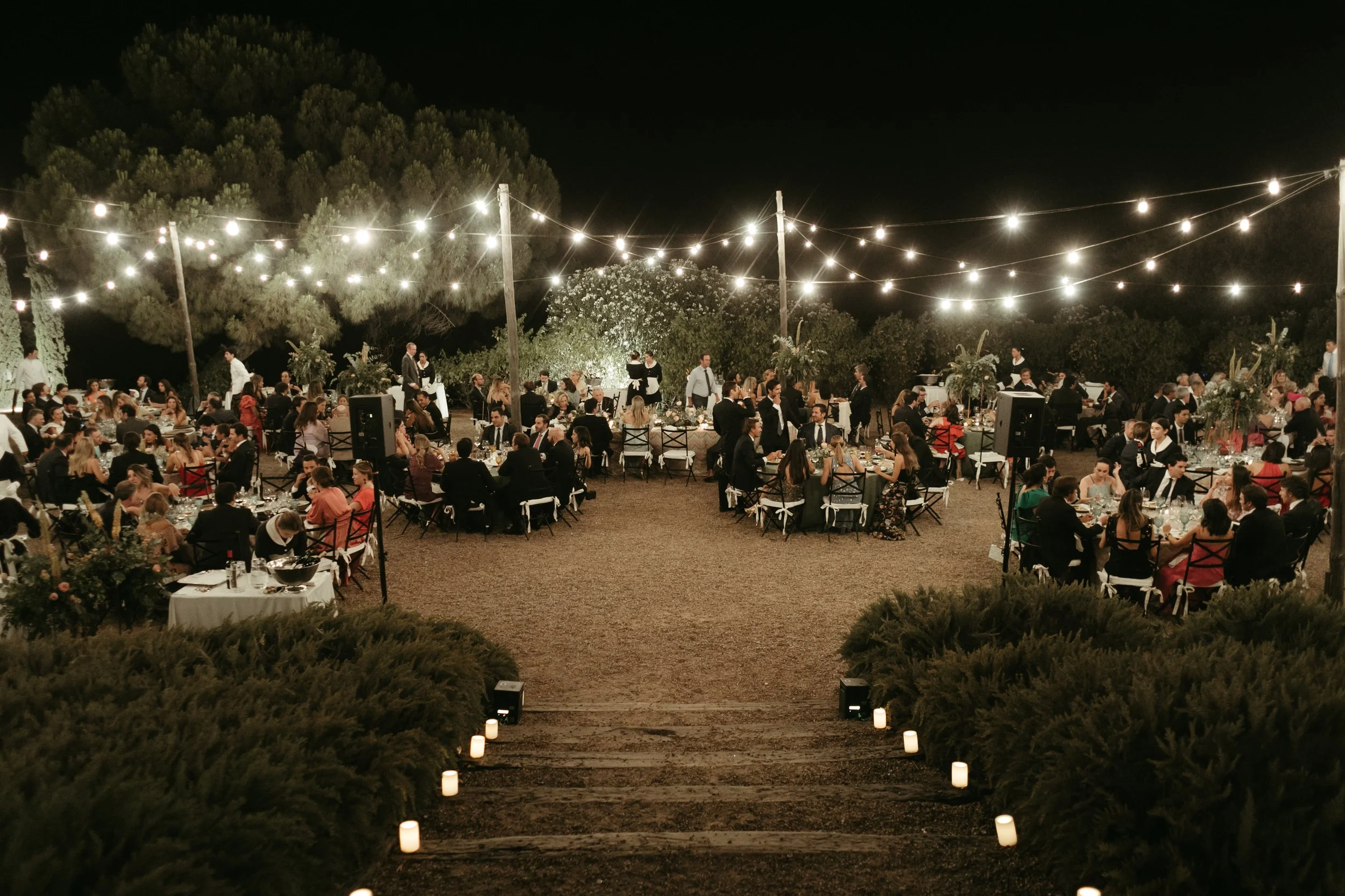 Celebración nocturna en un jardín con mesas decoradas y muchas personas, con luces colgantes y vegetación en el fondo.