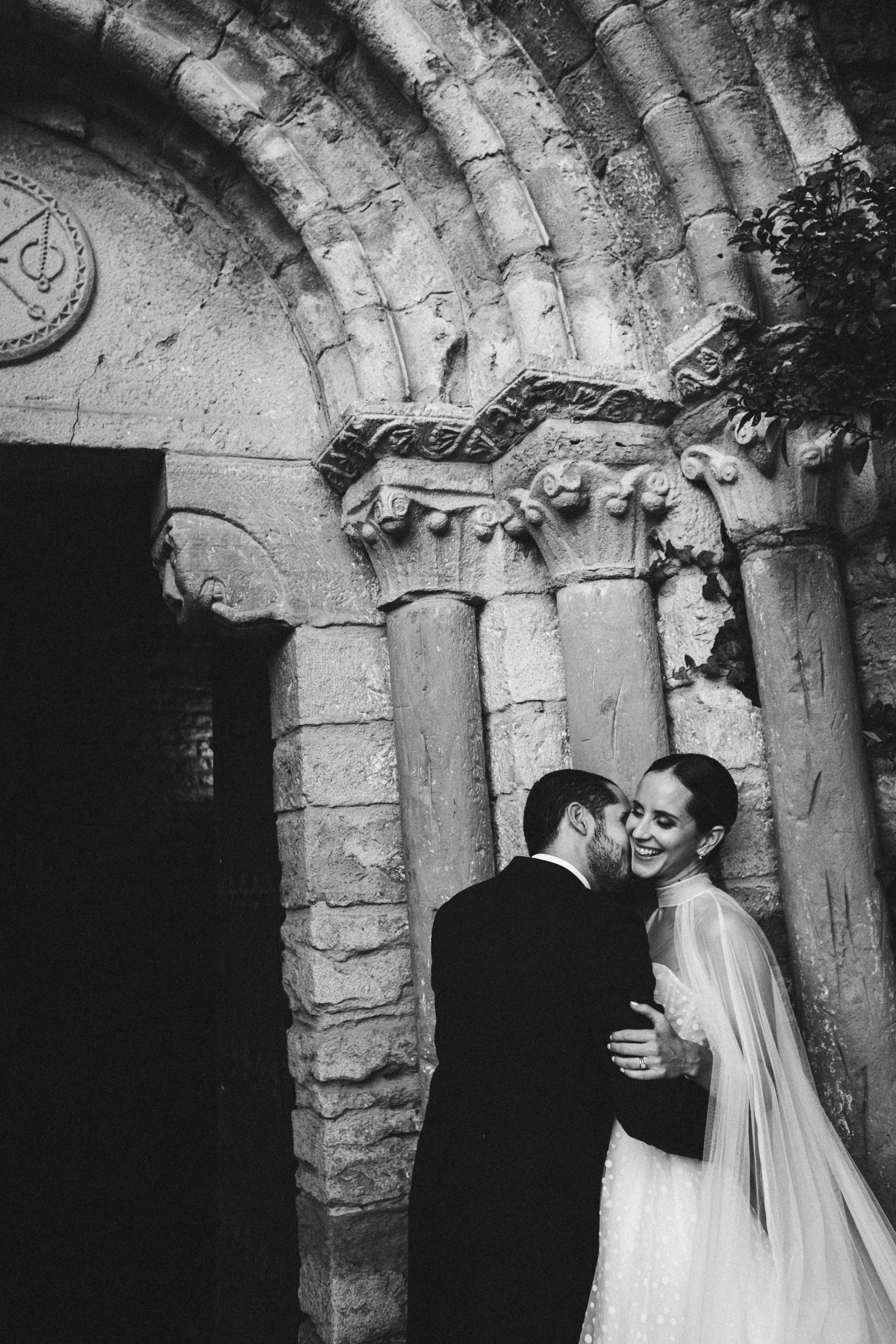 Pareja de novios en un fondo de arquitectura antigua de piedra, ella con vestido de novia y él con traje, sonriendo y abrazándose en positivo.