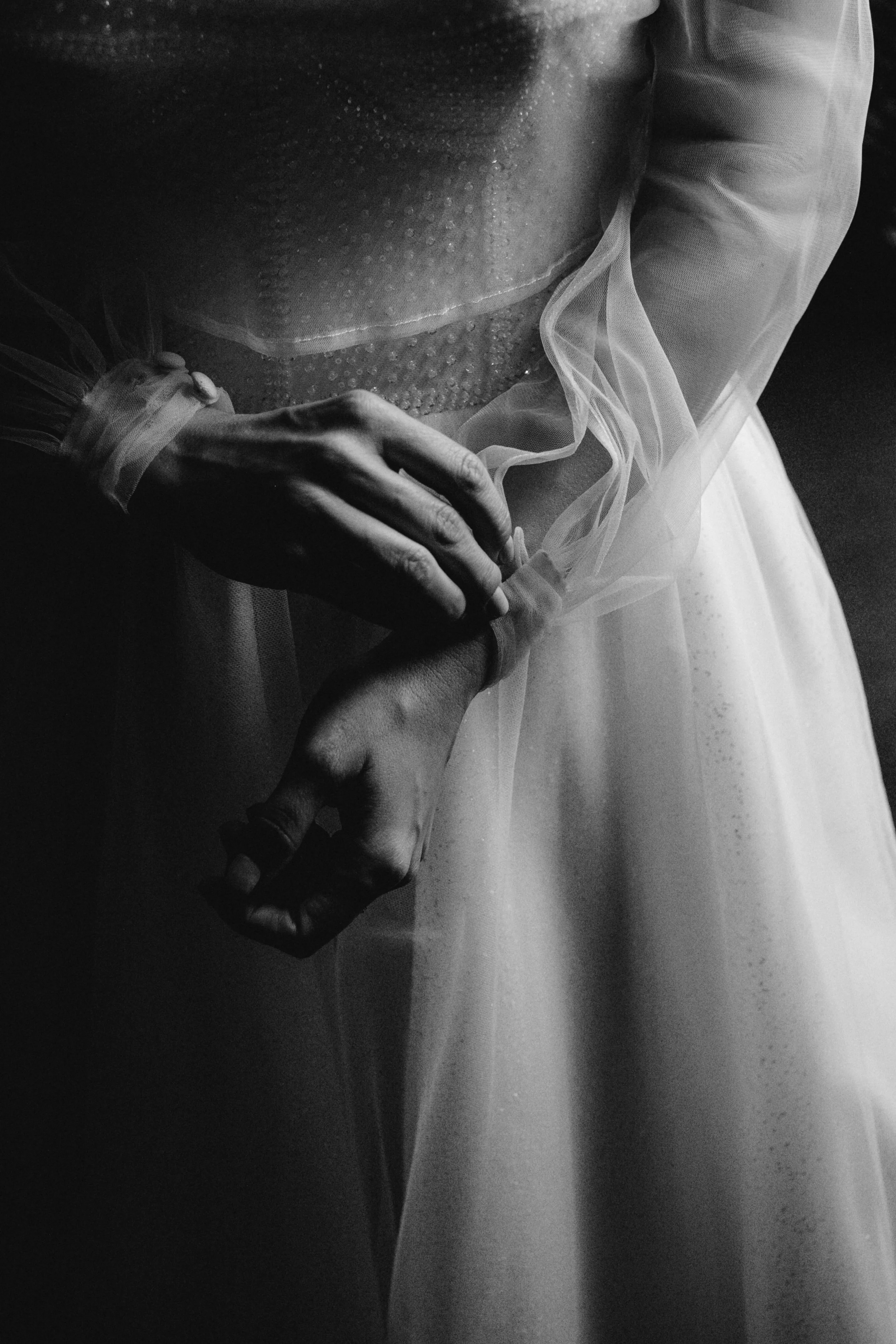 Primer plano en blanco y negro de unas manos entrelazadas, con vestido de gasa y detalles delicados, en un entorno oscuro.