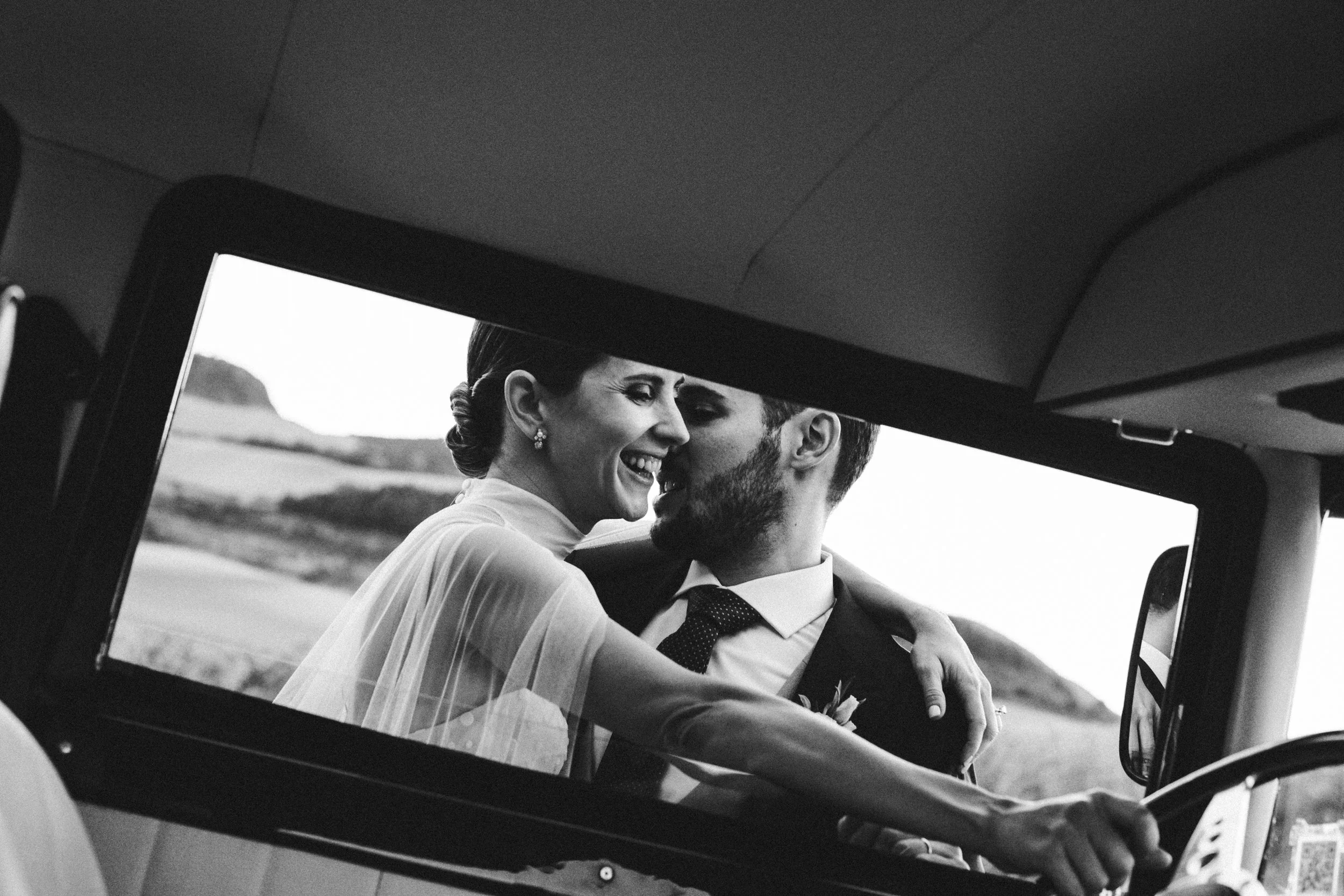Una pareja de recién casados sonriendo y abrazándose en el interior de un vehículo, con un fondo de paisaje rural.