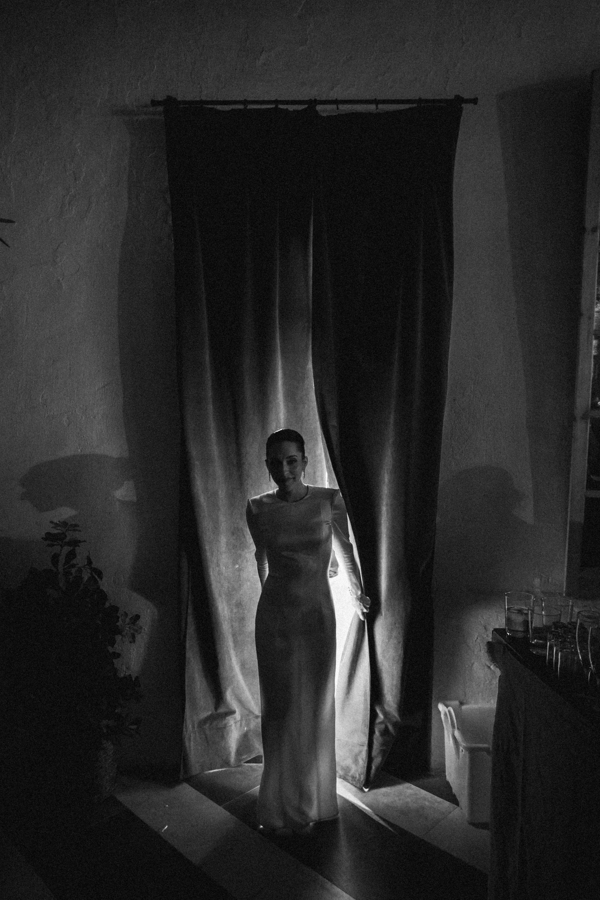 Mujer con vestido largo sale a través de cortina negra en una habitación con iluminación escasa, en tonos negros y blancos.