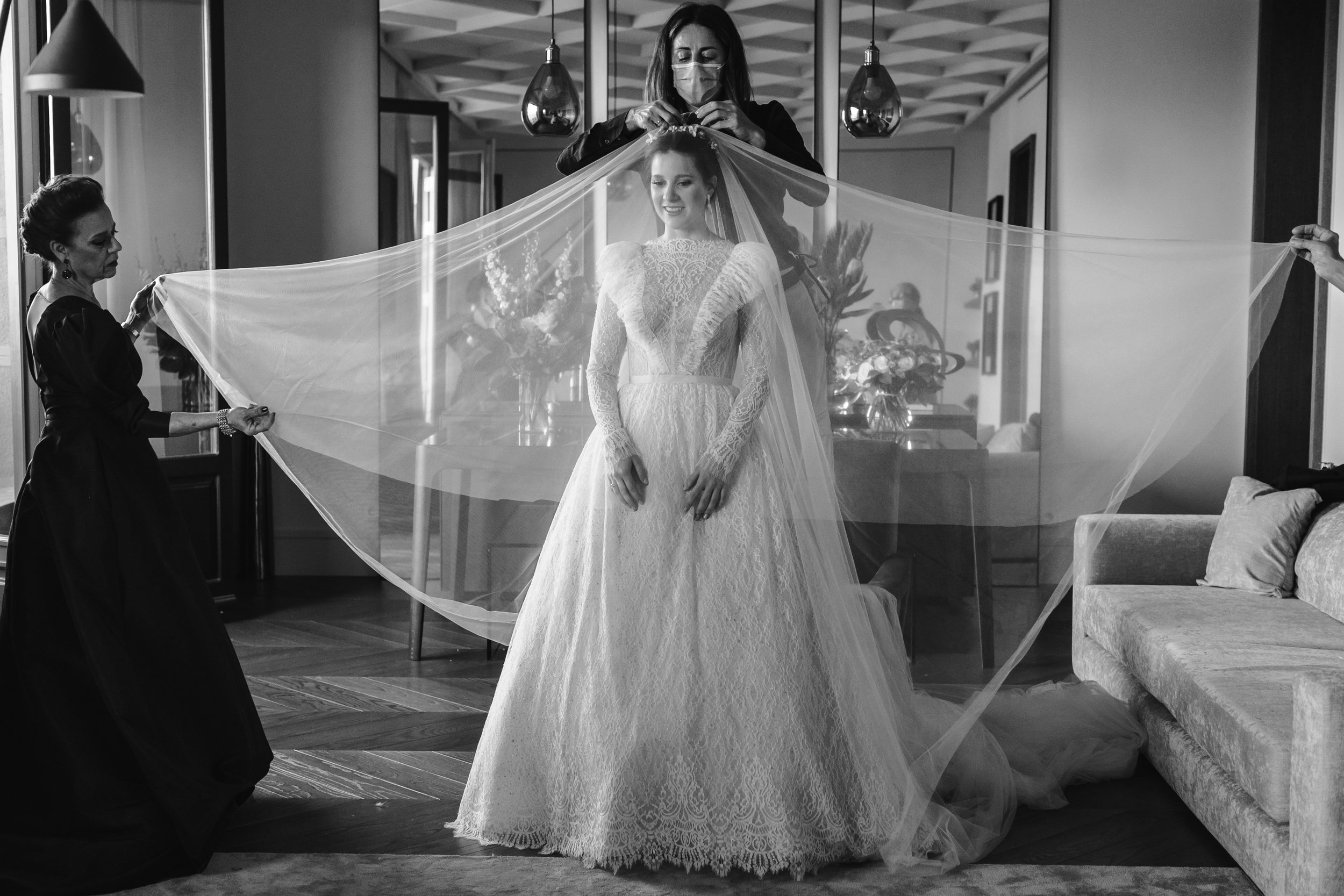 Una mujer vestida con un vestido de novia de encaje, en un interior, con varias personas ayudándola a colocarse su velo, en un ambiente elegante y decorado con flores.