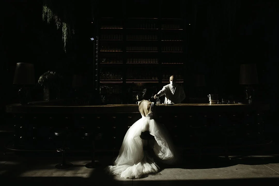 Una pareja en un bar, con la mujer de vestido de novia y el hombre de espalda, en un ambiente oscuro y elegante.