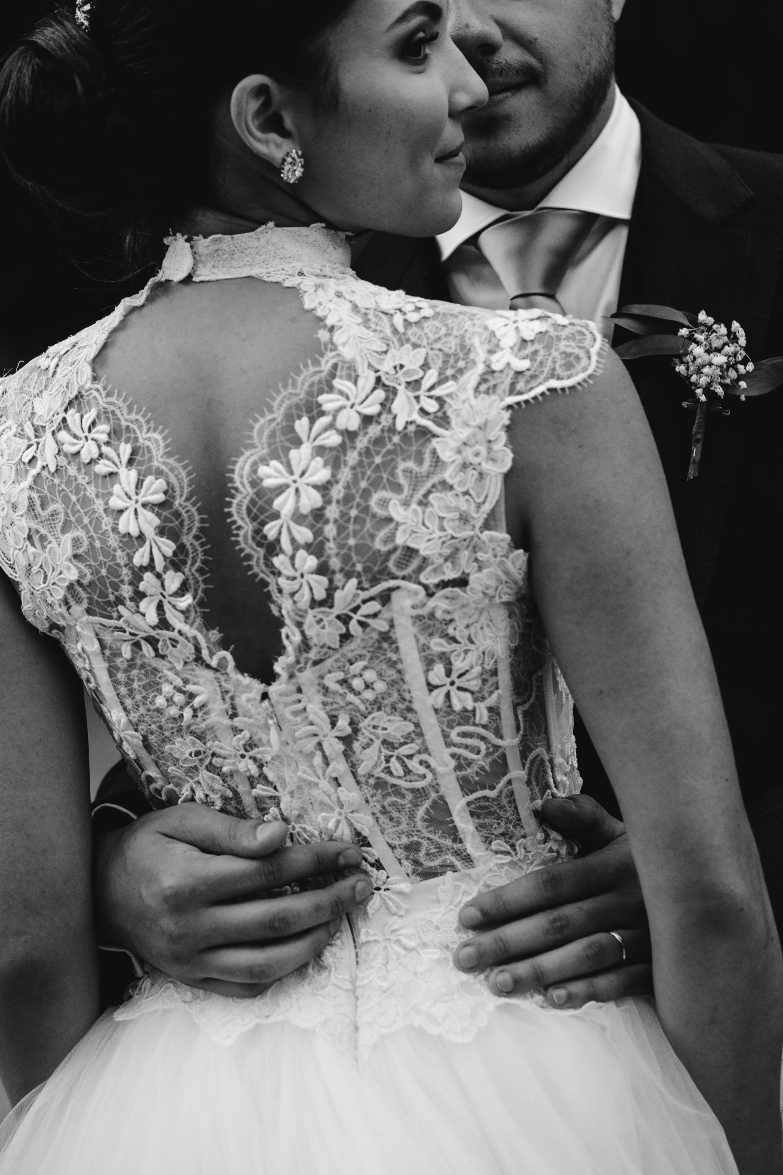 Pareja de boda en un abrazo, la mujer viste un vestido de encaje con detalles florales y el hombre lleva un traje con corbata y flor en el saco.