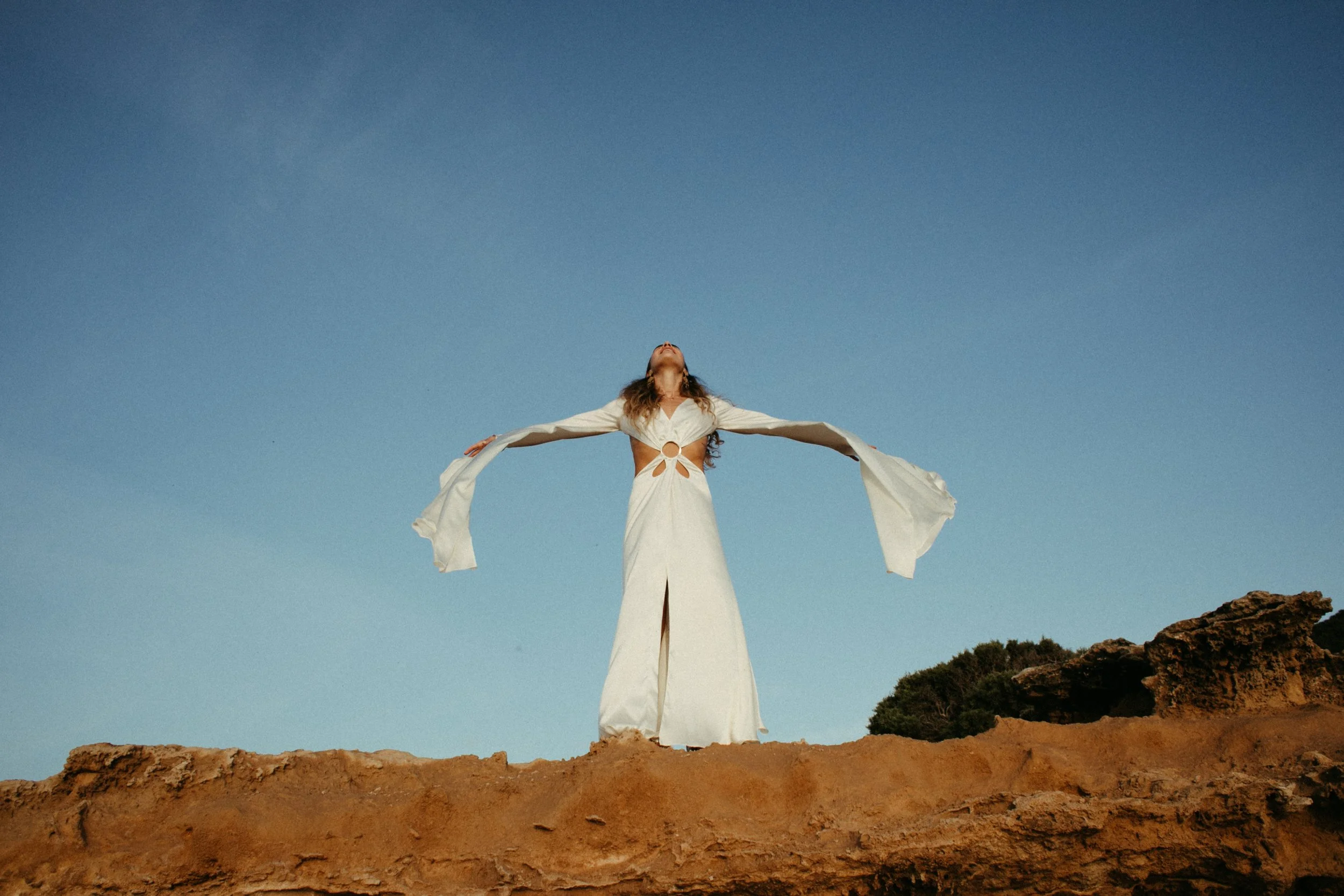 Mujer con vestido blanco de pie en una roca con los brazos extendidos y la cabeza inclinada hacia atrás, bajo un cielo despejado.