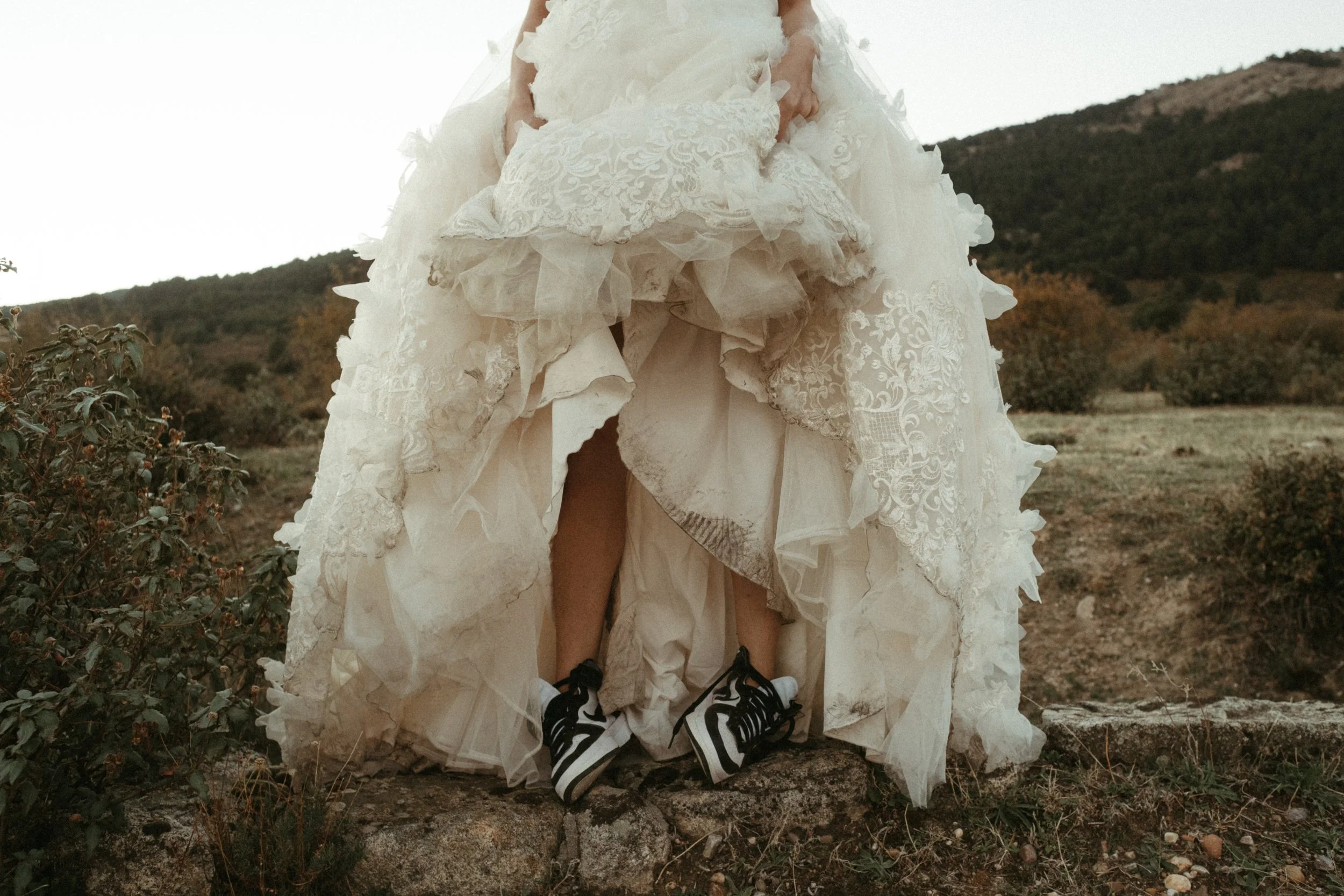 Persona con vestido de novia y zapatos deportivos, levantando la falda en un paisaje al aire libre con colinas y arbustos.