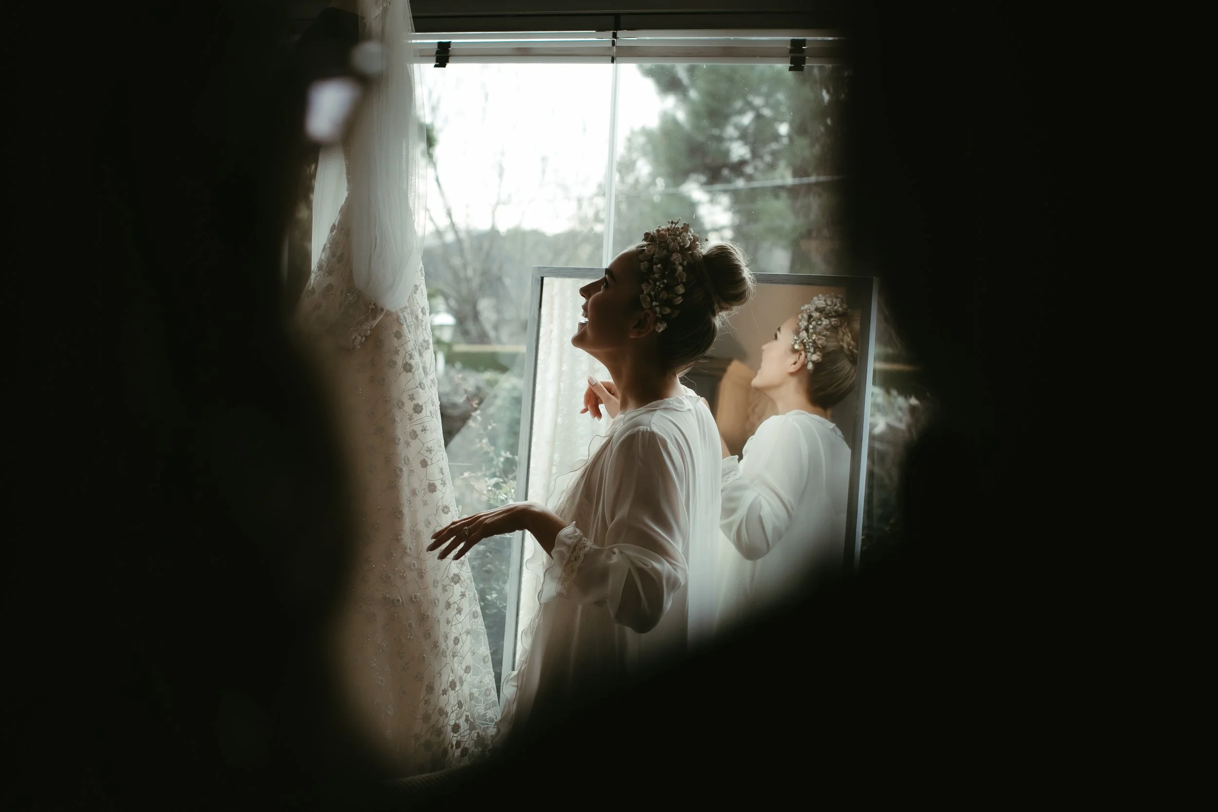 Mujer con peinado con flores, vestida con bata, mirándose en un espejo, en un ambiente luminoso con ventana y cortinas, probablemente preparándose para una ocasión especial.