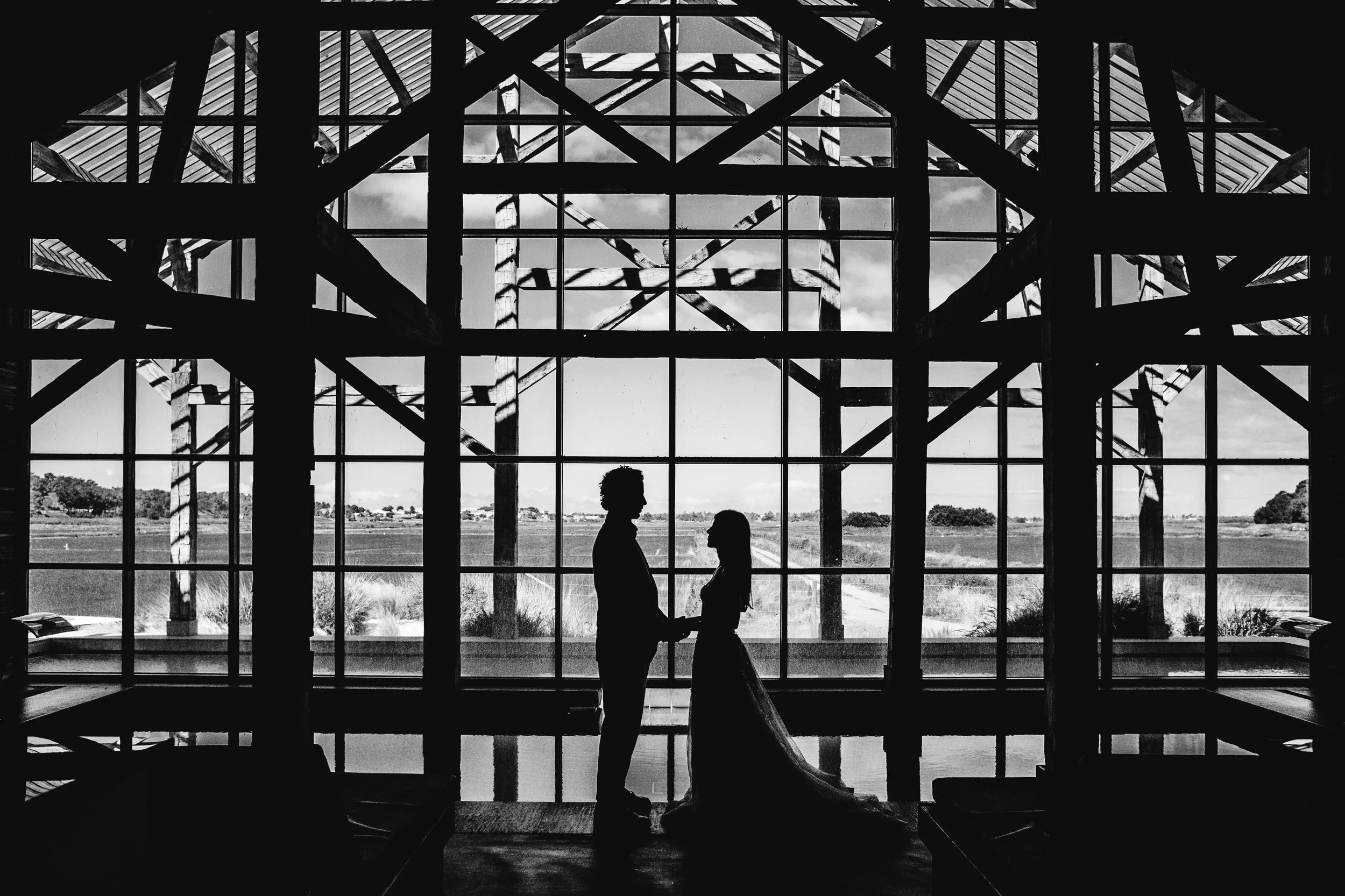 Pareja de novios en silueta sosteniéndose de las manos frente a una gran ventana con estructura de madera y vidrio, en un espacio con vista a un paisaje rural con campos y árboles.