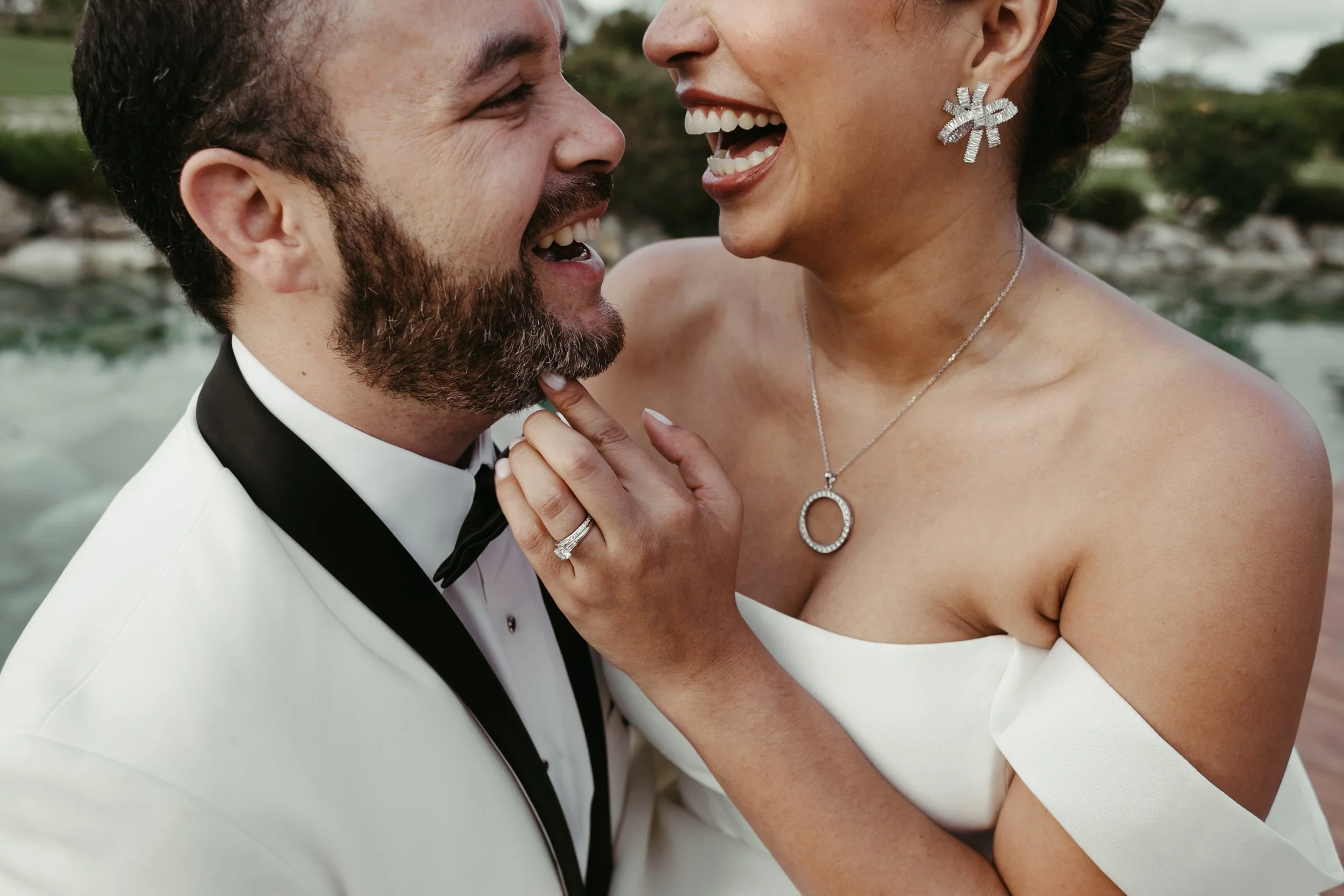 Pareja de recién casados riendo, en boda, con fondo natural de agua y árboles.