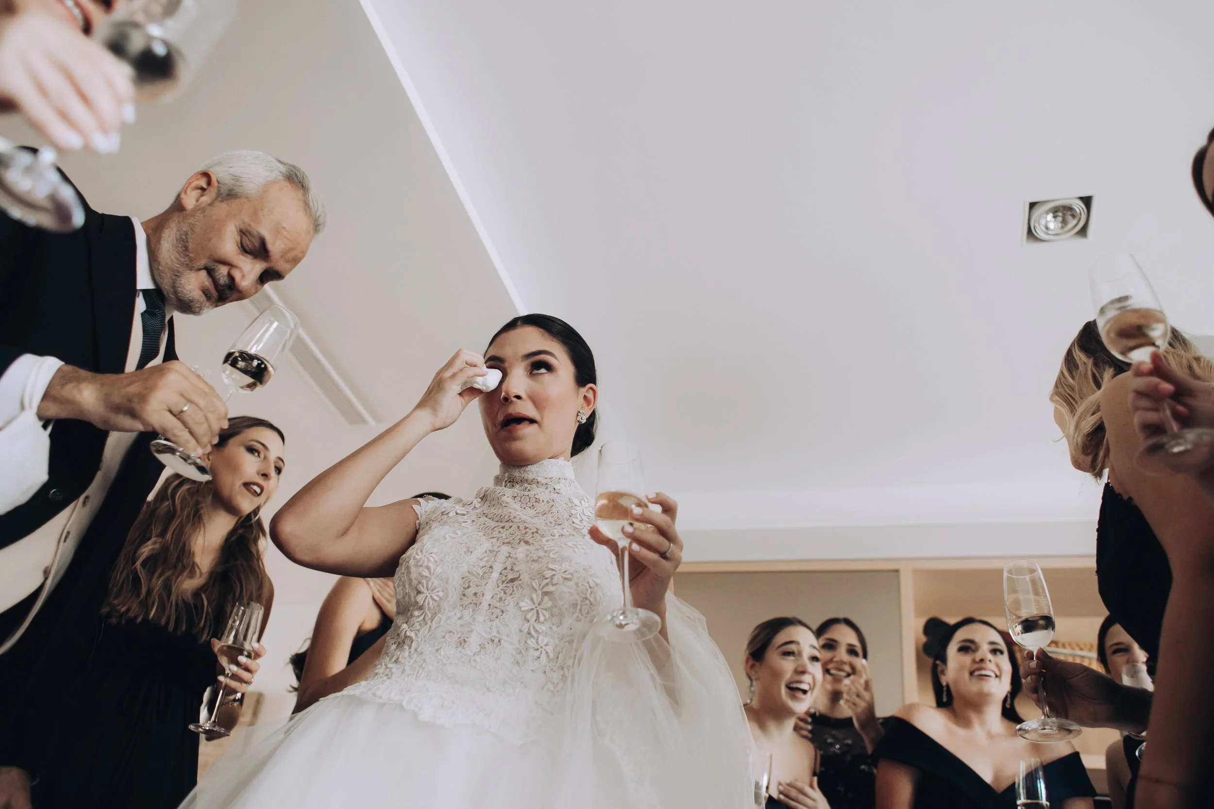 Mujer en vestido de novia llorando y sus amigos en celebración con copas de vino en una reunión social.
