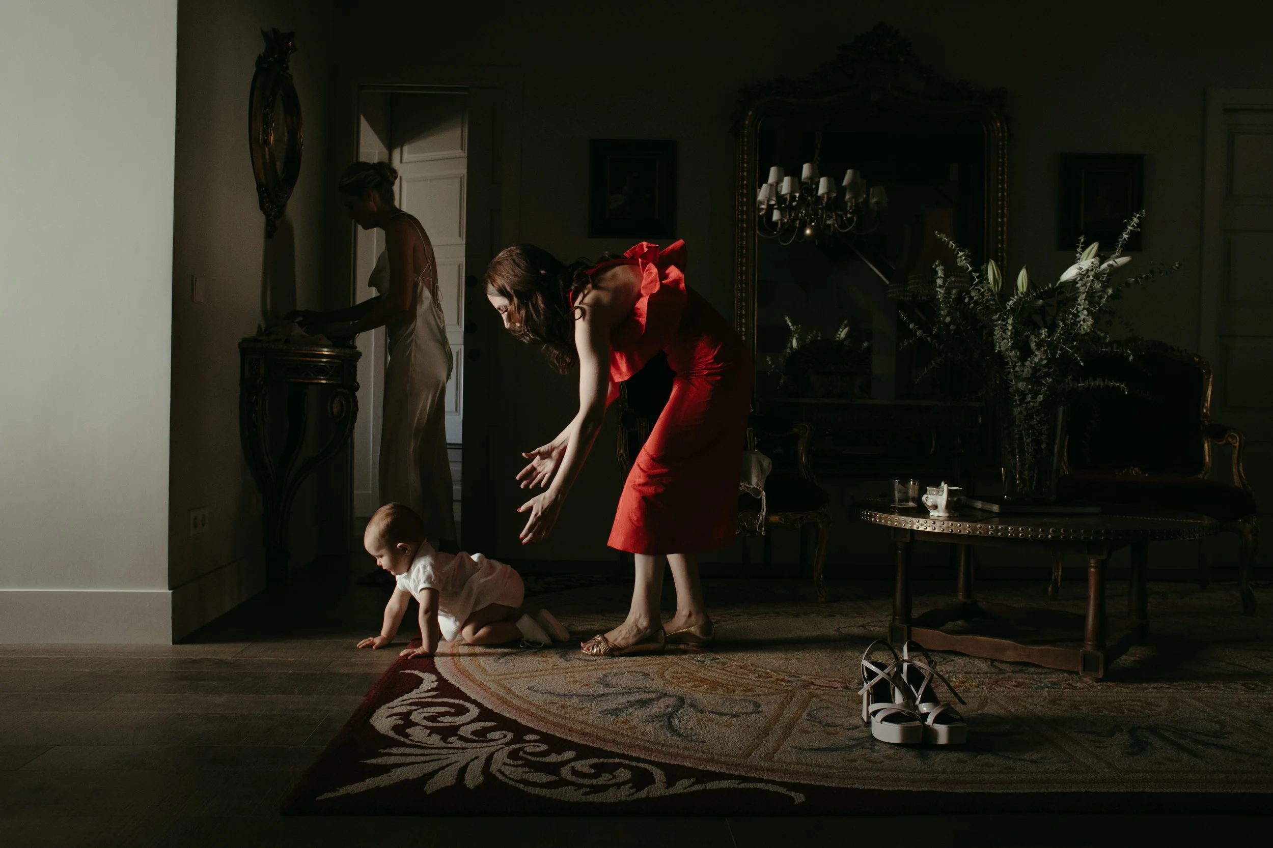 Mujer en vestido rojo doblándose para recoger un niño que gatea sobre una alfombra en un salón oscuro, otra mujer en vestido blanco en el fondo junto a una mesa decorada con flores y objetos.