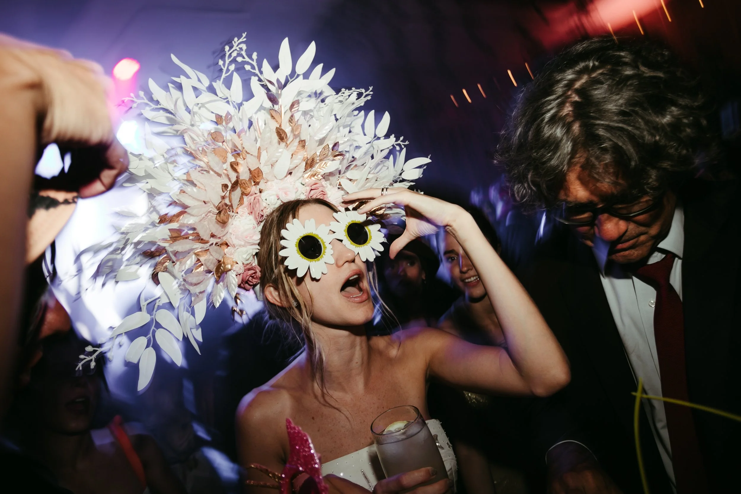 Mujer con gafas de sol con diseño de margaritas, vestido blanco, en una fiesta con un sombrero de flores y hojas y varias personas bailando y disfrutando la celebración.