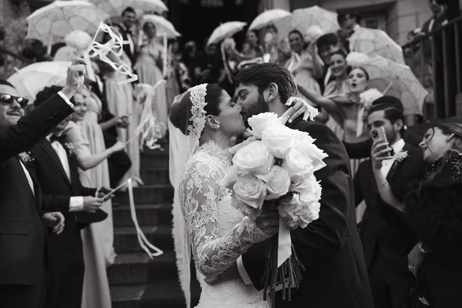 Black and white photo of the newlyweds kissing as guests wave ribbon wands outside the church, Madrid