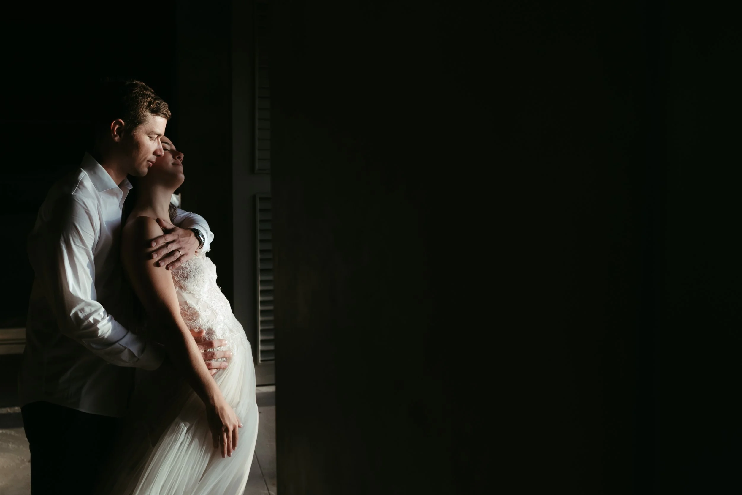 Pareja abrazada y con los ojos cerrados frente a una ventana con luz tenue.