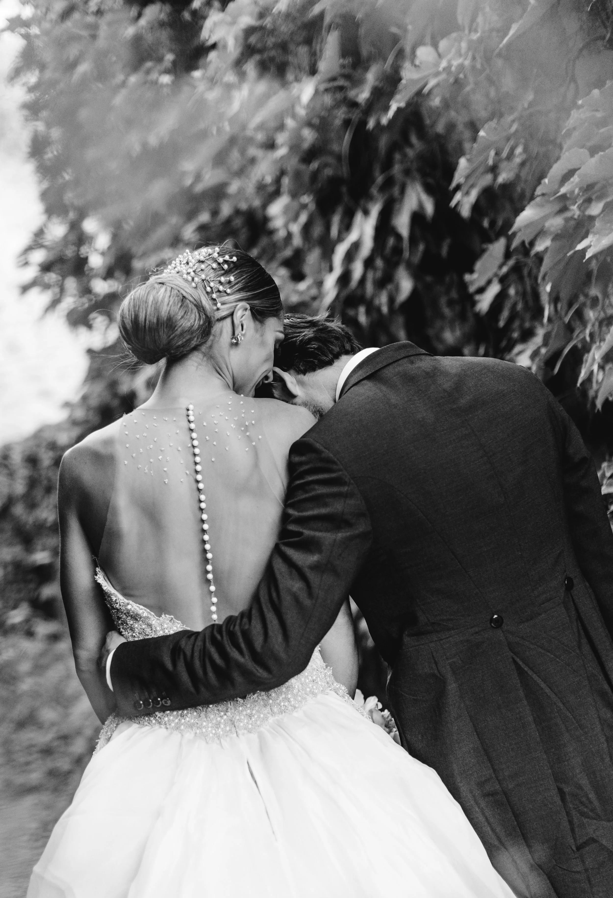 Pareja de novios en boda, abrazados y risueños, al aire libre rodeados de hojas.