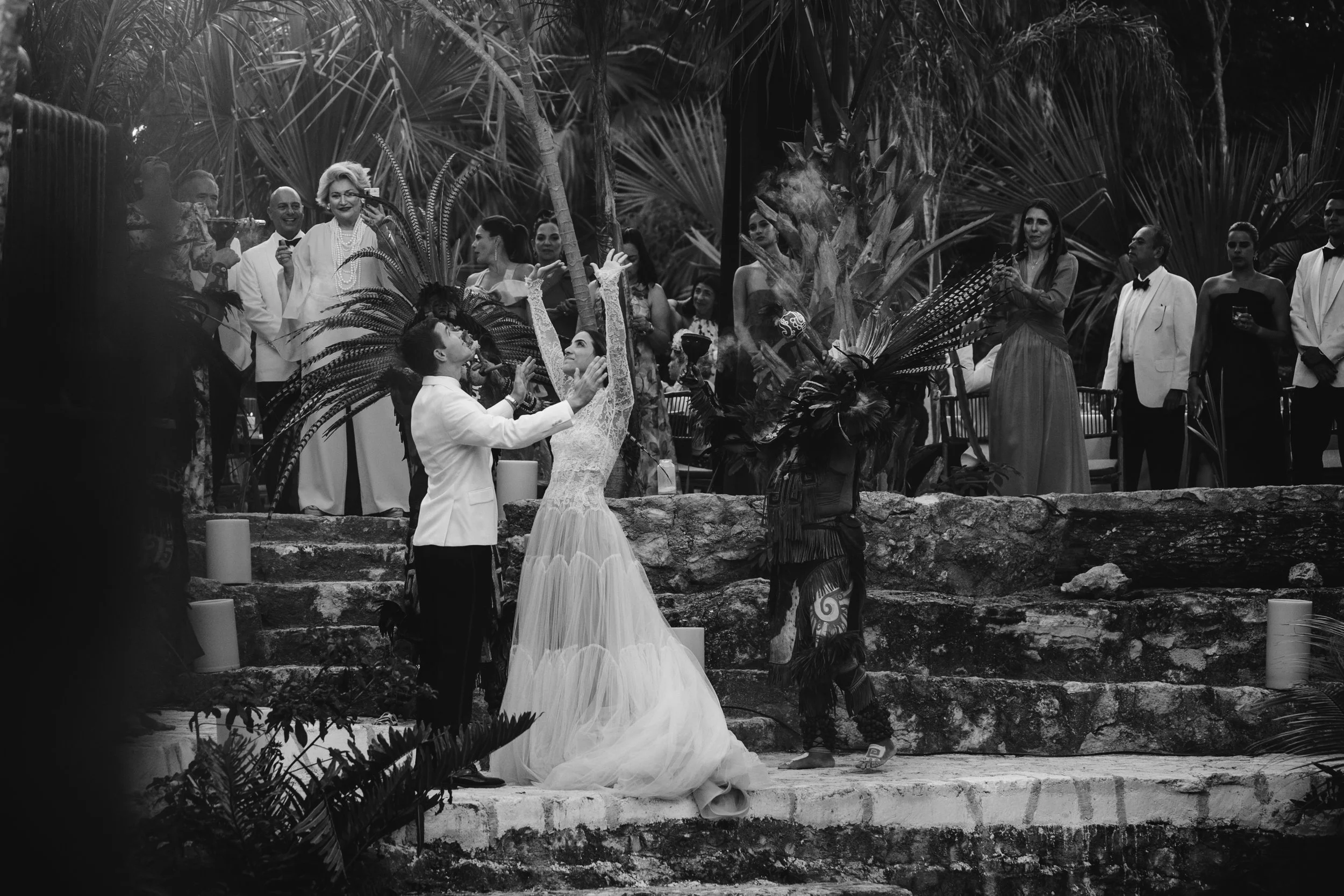 Una boda con ceremonia en un entorno tropical, con personas vestidas elegantemente, decoraciones con plumas y velas.