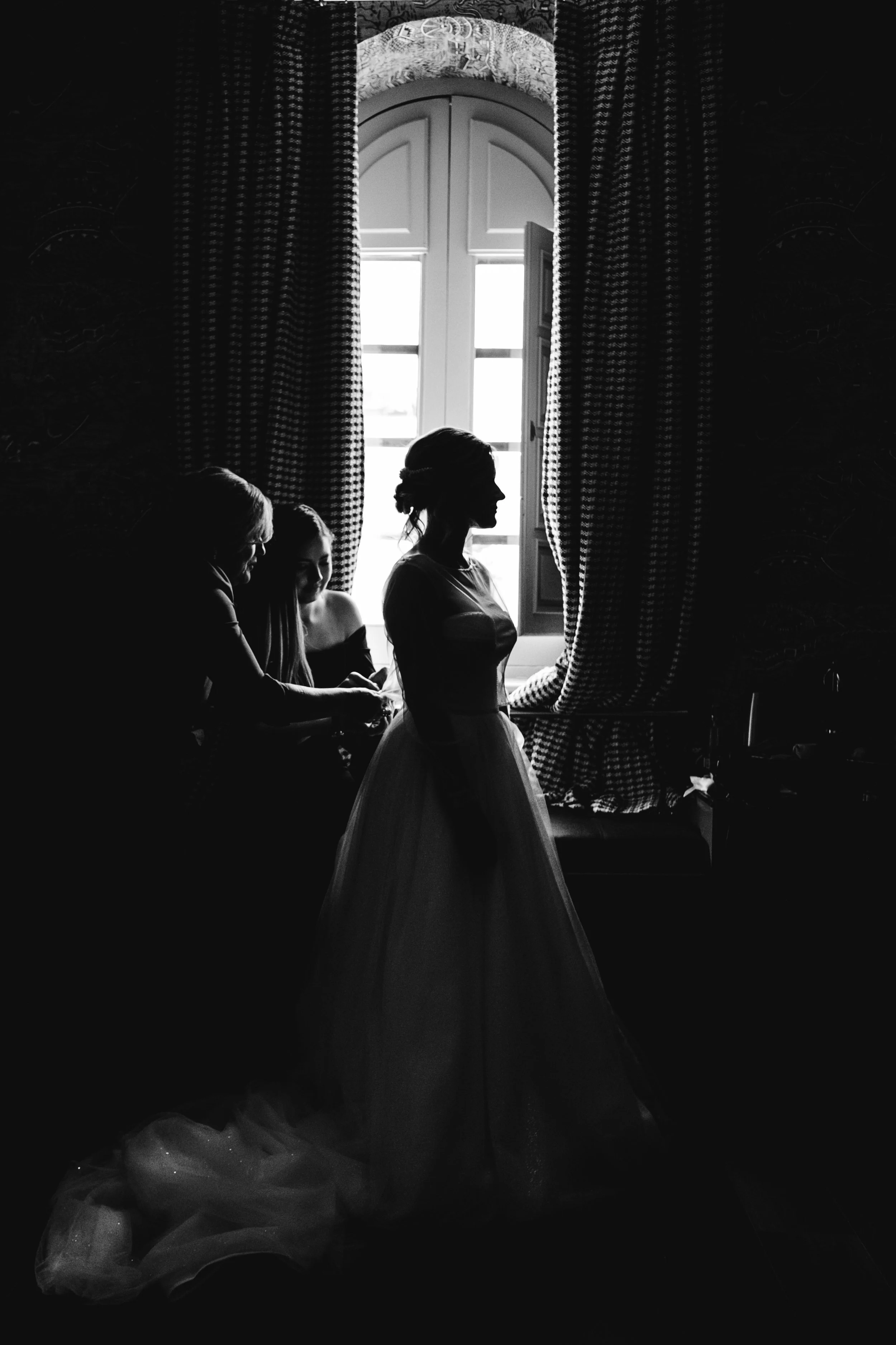 Mujer con vestido largo de novia en silencio en una habitación, con otras tres mujeres sentadas junto a ella, frente a una ventana con cortinas de cuadros y luz que ilumina desde atrás.
