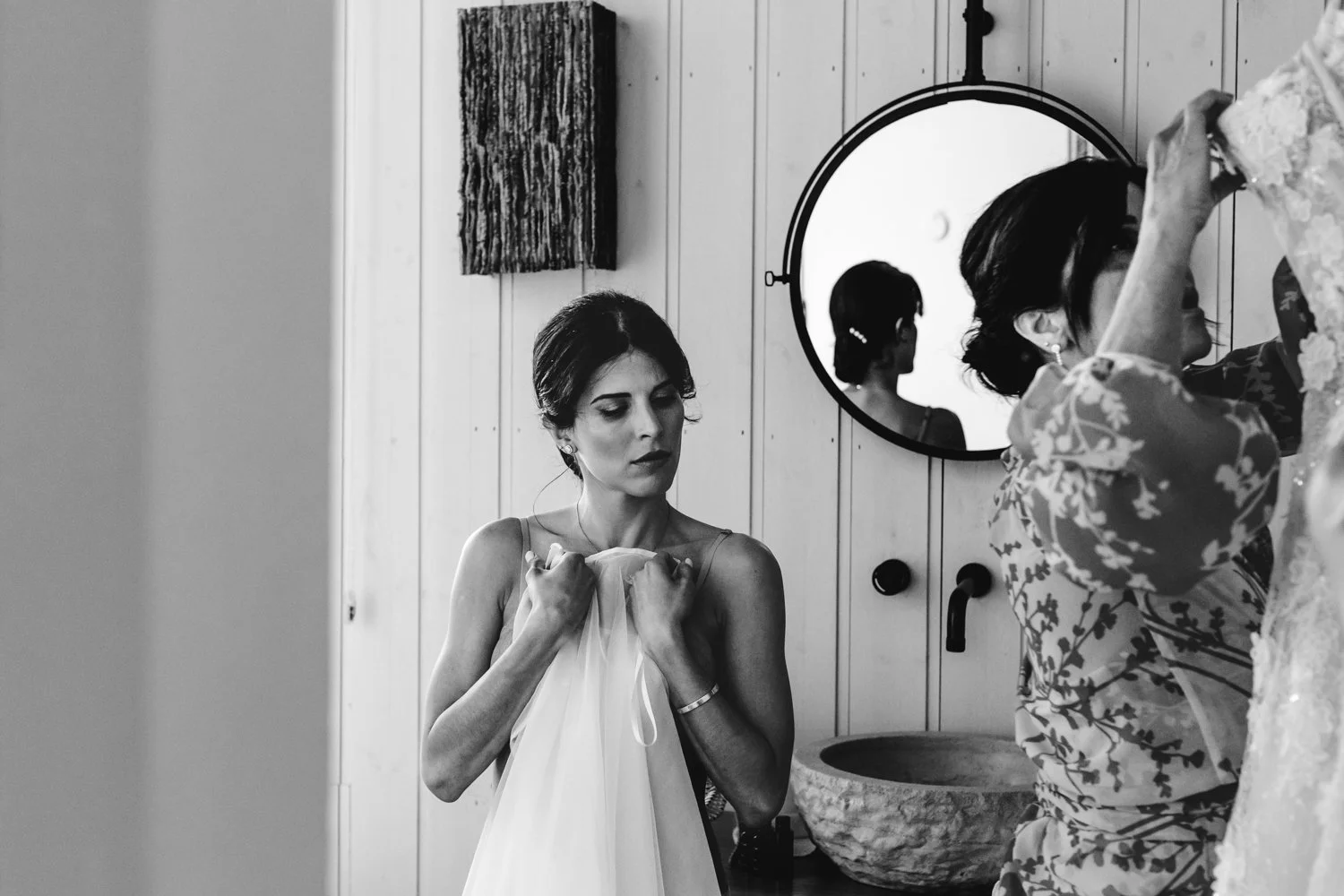 Mujer joven con vestido de novia ajustándose la ropa en un ambiente con paredes de madera y espejos redondos.
