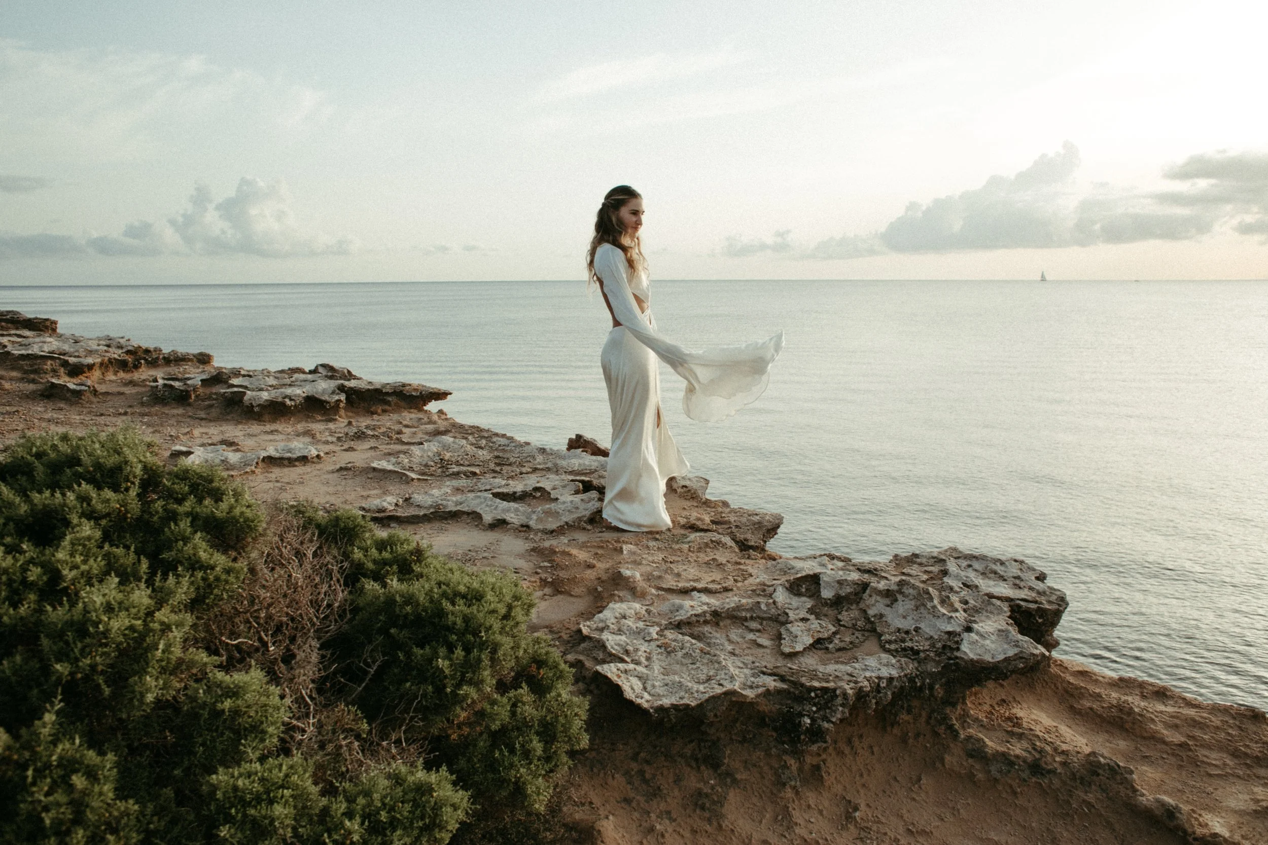 Mujer con vestido blanco en la orilla rocosa del mar en atardecer, vista panorámica del océano y cielo nublado en el fondo.