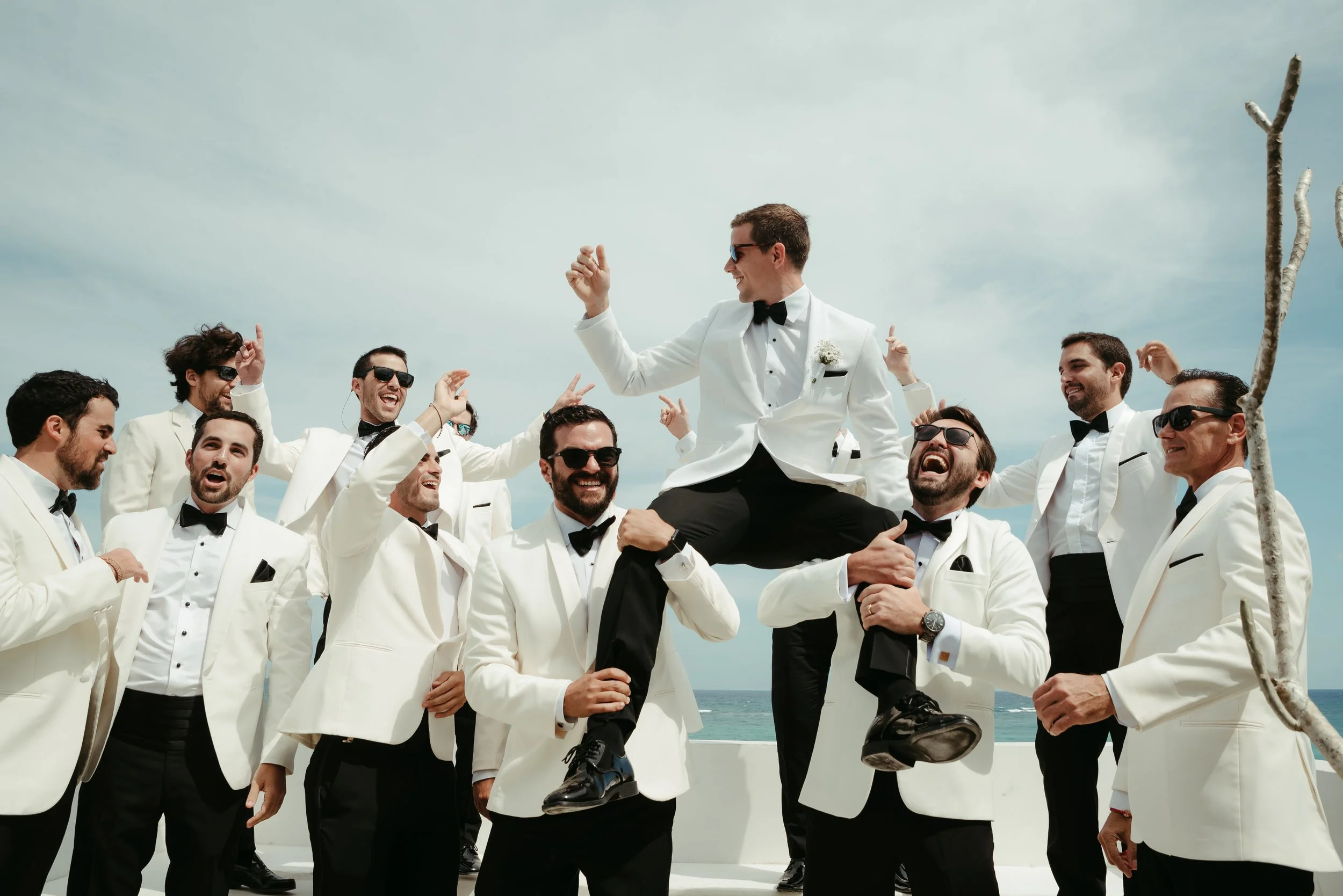 Grupo de hombres en traje blanco y corbata de lazo, celebrando en la playa, con uno de ellos en brazos del otro mientras lo levantan, todos sonriendo y usando gafas de sol.