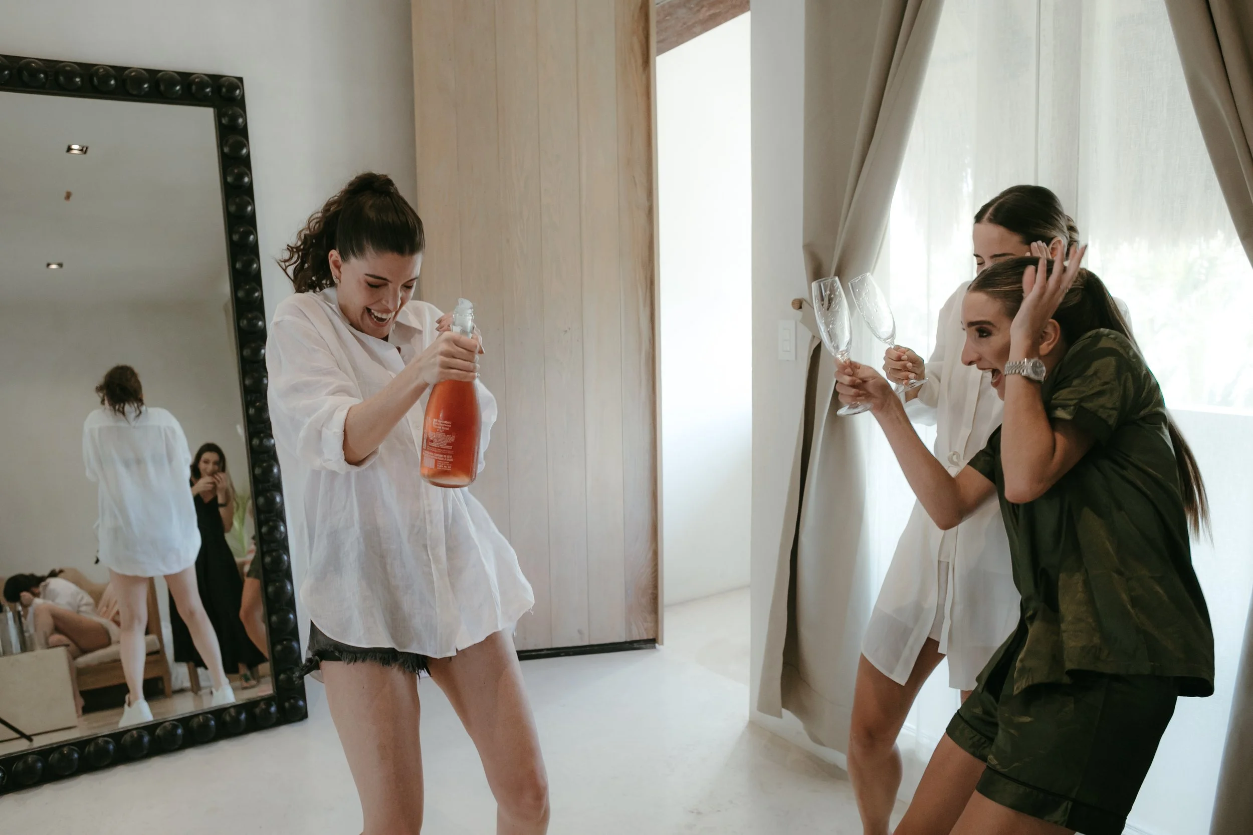 Grupo de mujeres celebrando con risas y brindis en un ambiente relajado, una de ellas sosteniendo una botella de champán y las otras con copas de vino.