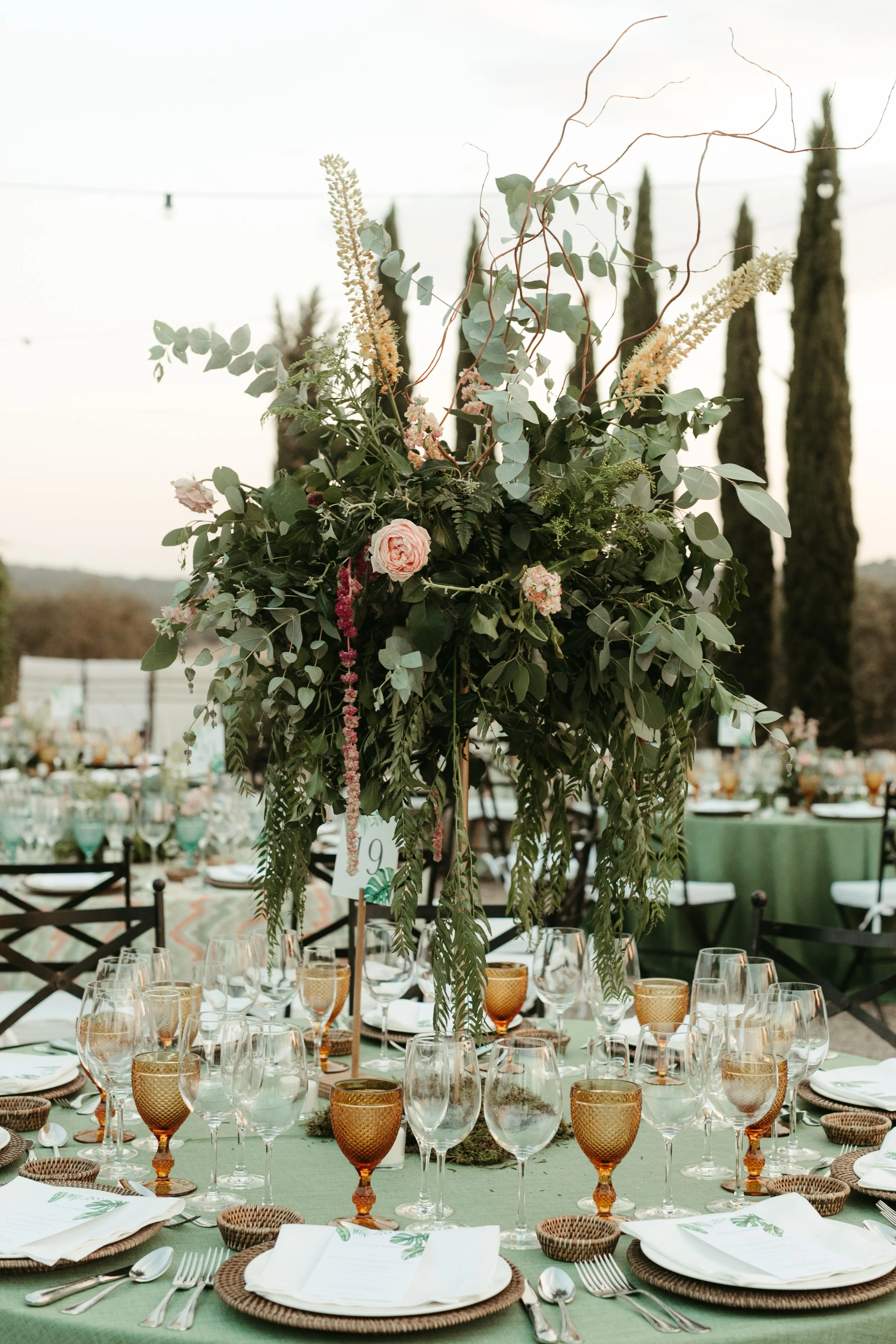 Mesa decorada para evento al aire libre con centro de flores y hojas verdes, vasos de vino y copas en tonos dorados, platos y cubiertos ordenados, manteles verdes y sillas negras.