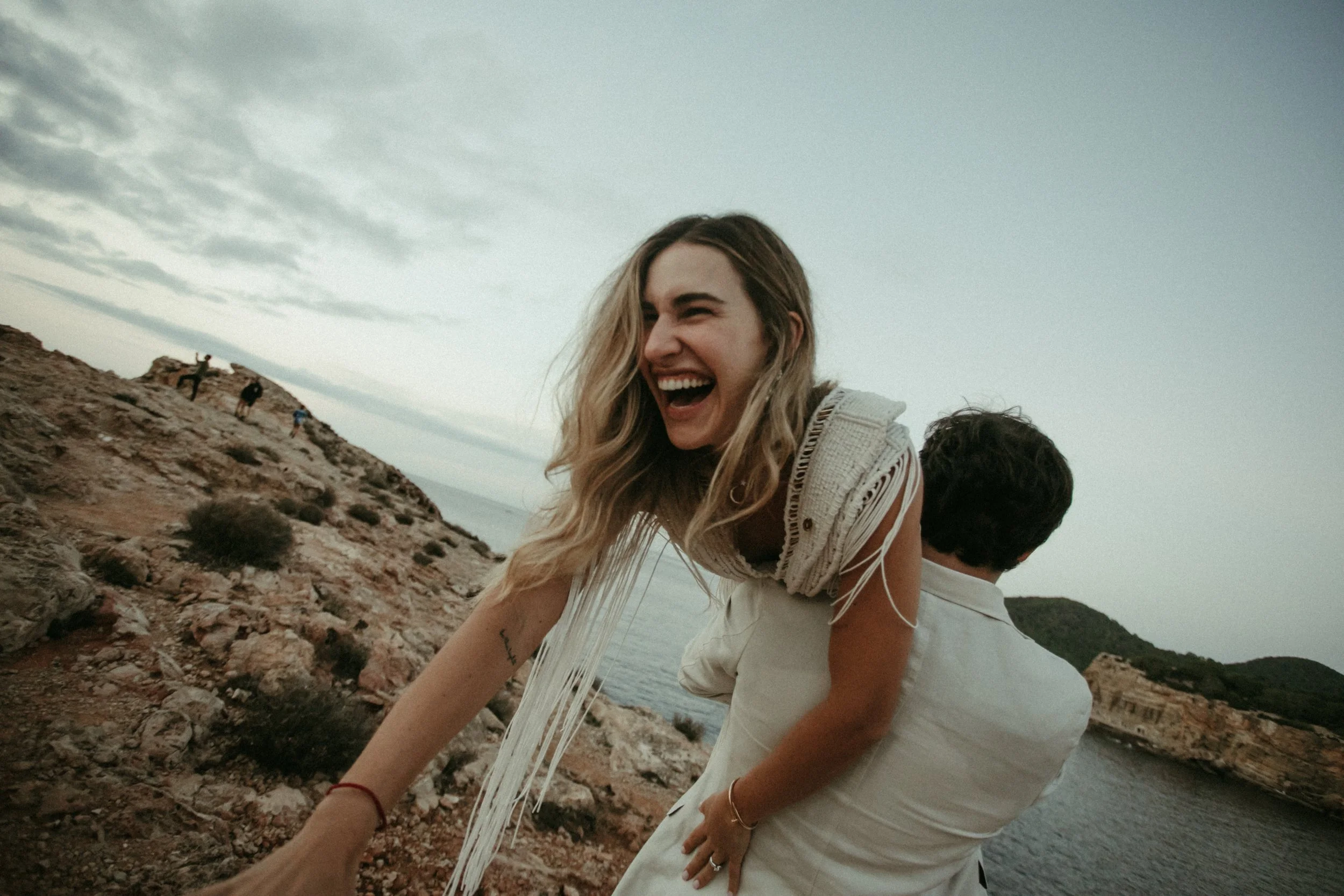 Una pareja disfrutando en la playa, la mujer ríe mientras está en los brazos del hombre, con un fondo de cielo nublado, montaña y mar.