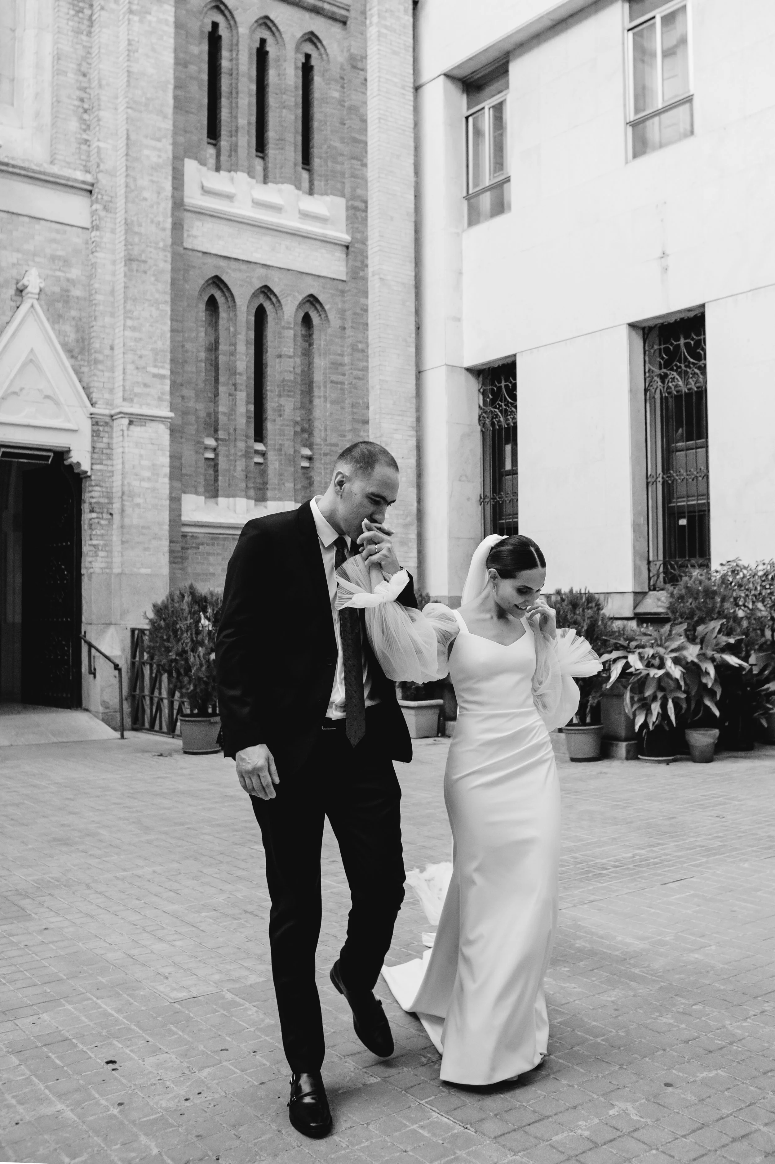 Pareja de novios caminando juntos afuera de un edificio, la novia lleva vestido largo y velo, el novio usa traje negro.