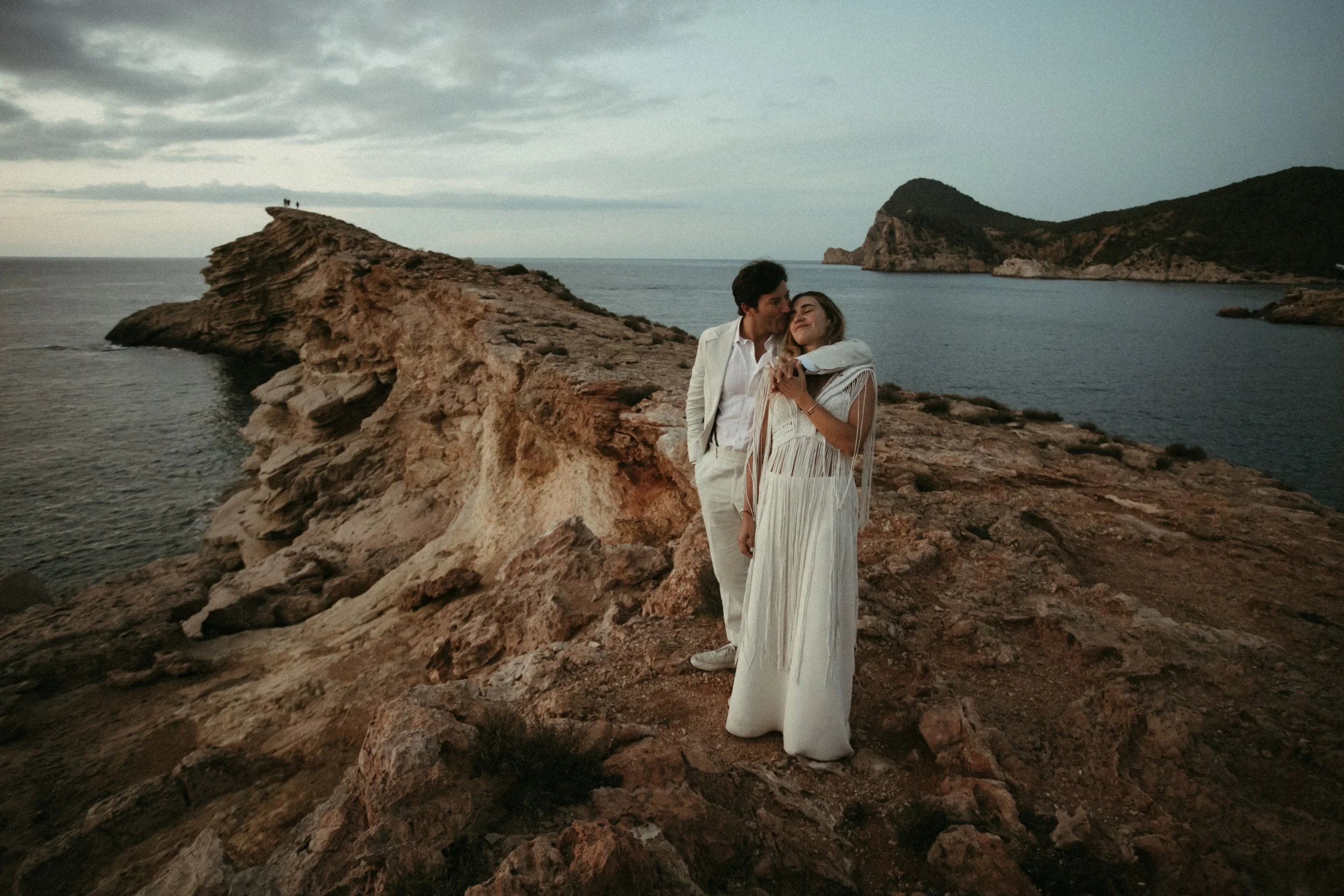 Una pareja en un vestido y traje blanco en un paisaje costero con rocas y mar al atardecer.