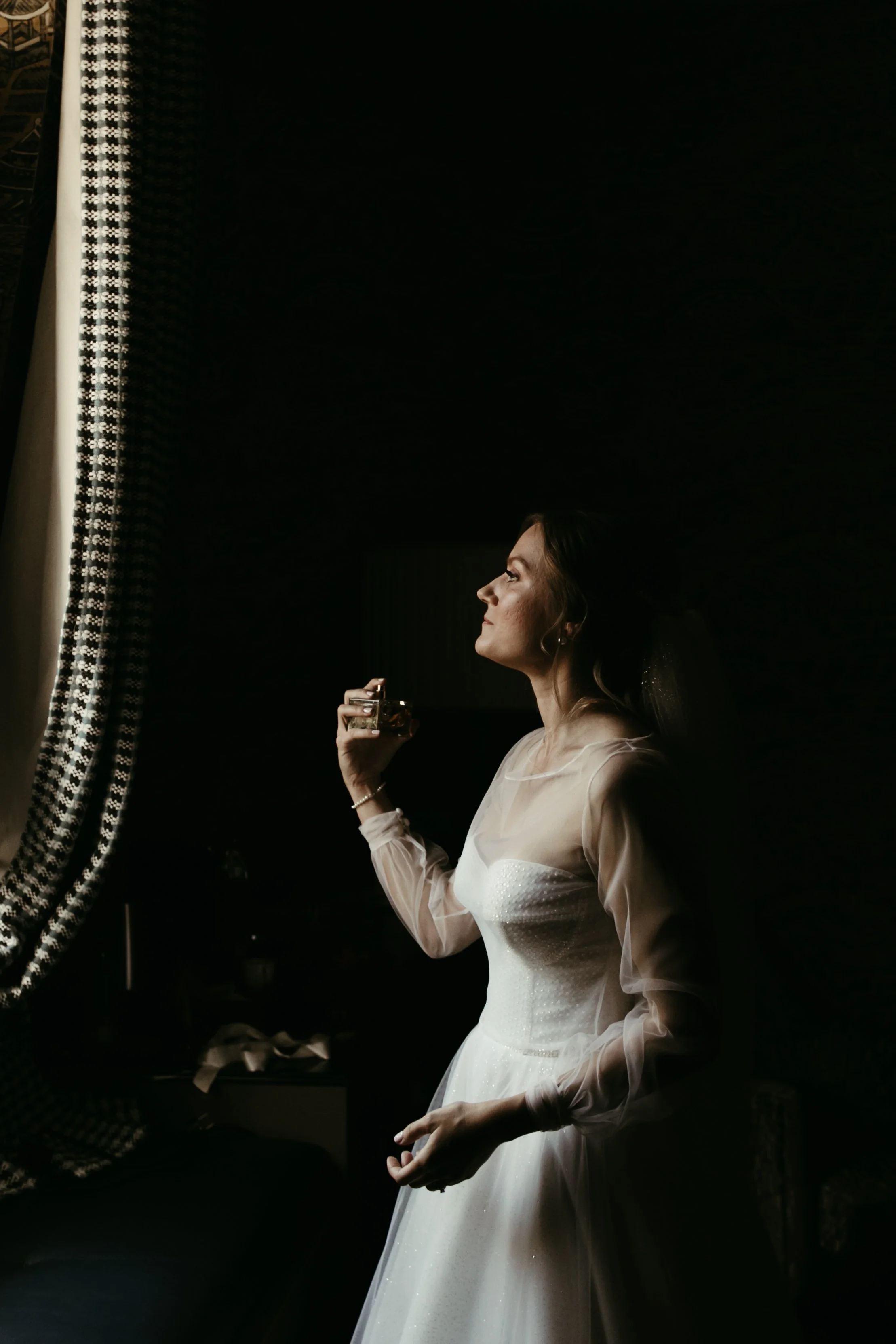Una mujer vestida con un vestido blanco, probablemente una novia, sosteniendo un vaso en una mano y mirando hacia una ventana con cortinas de ganchillo, en un ambiente oscuro con luz natural entrando.